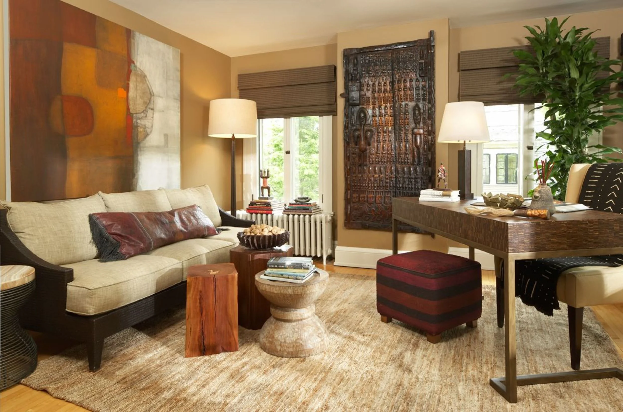 Living room with beige sofa, wooden side tables, stack of books, large abstract painting, tribal art piece on wall, tall lamps, windows with brown shades, green plant, desk with decorative items, and a textured rug.