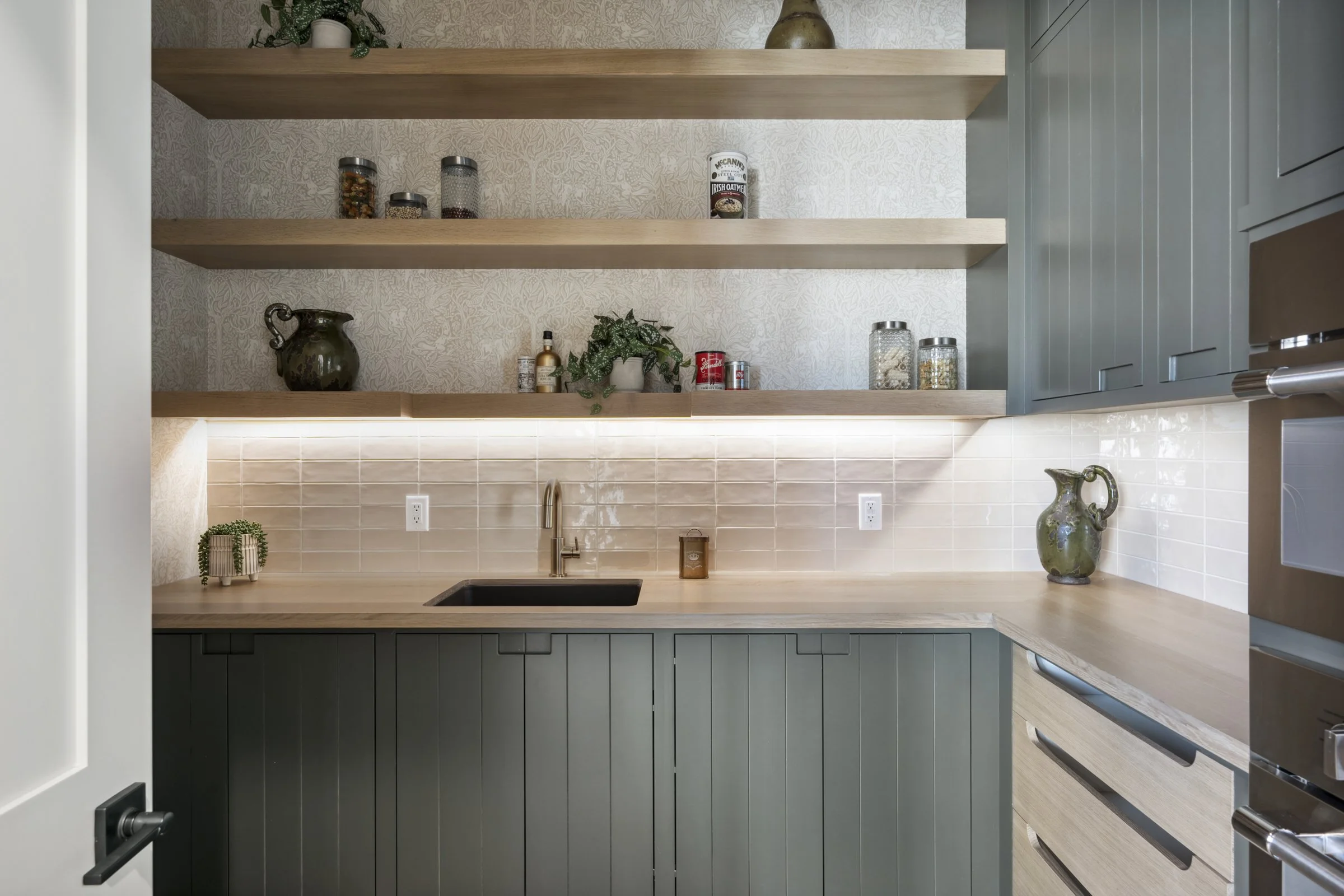 Modern kitchen with light wood countertops, dark green lower cabinets, and open shelves with jars, plants, and decorative items.