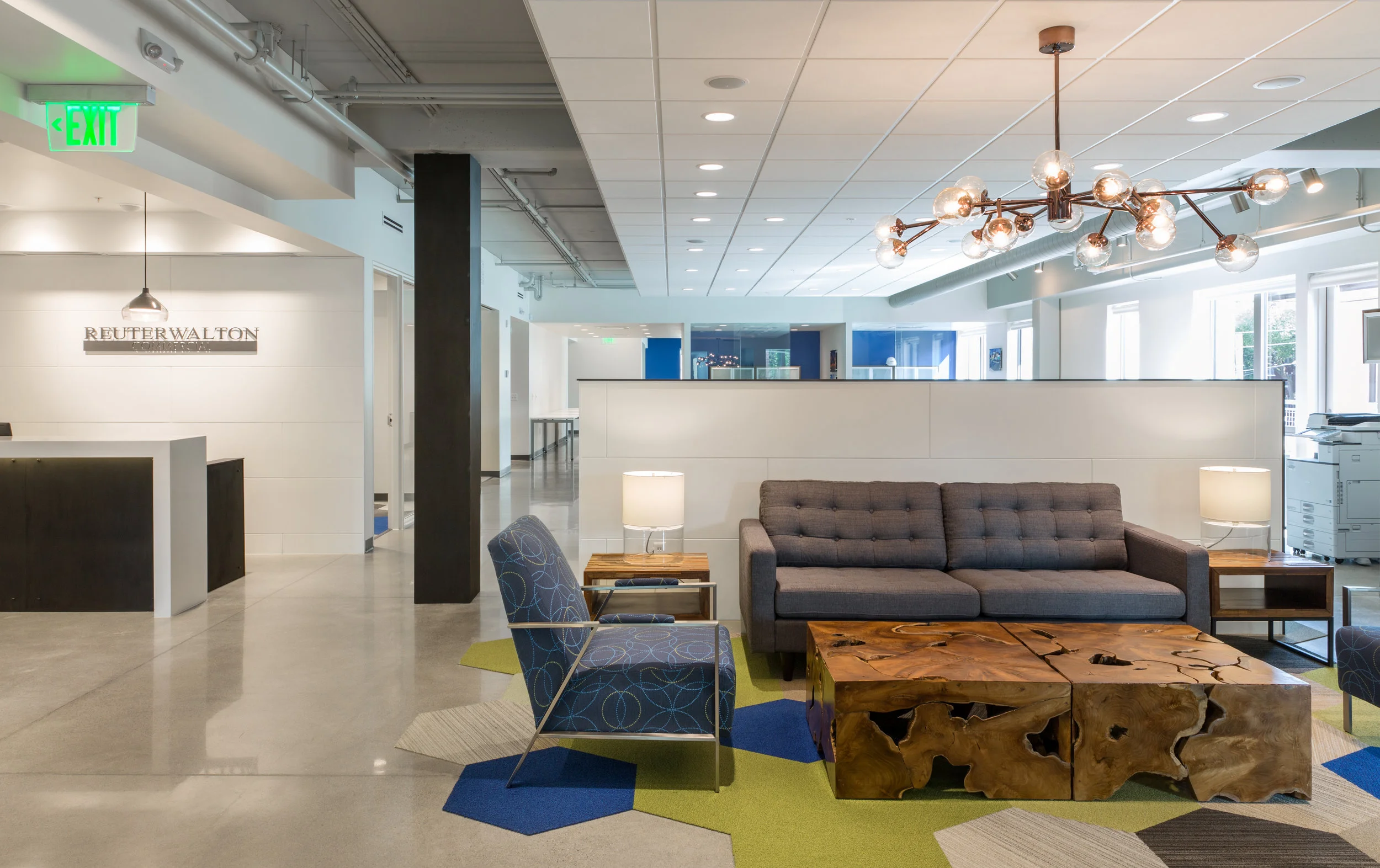 Modern office reception area with gray couch, armchair, wooden coffee table, lamps, and contemporary chandelier, with white walls, large windows, and blue accent wall in the background.