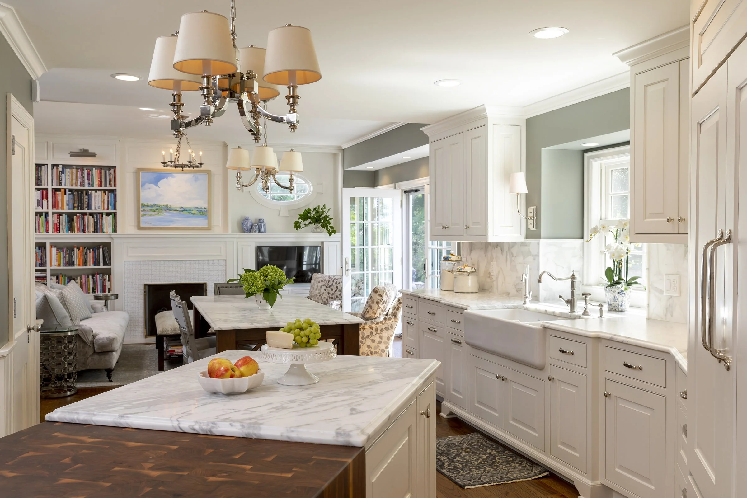 Bright kitchen with white cabinetry, marble countertops, a farmhouse sink, and natural light from windows. A chandelier hangs above a marble island, which has apples, grapes, and a cake stand with cheese. Past the kitchen, a cozy living area with a b