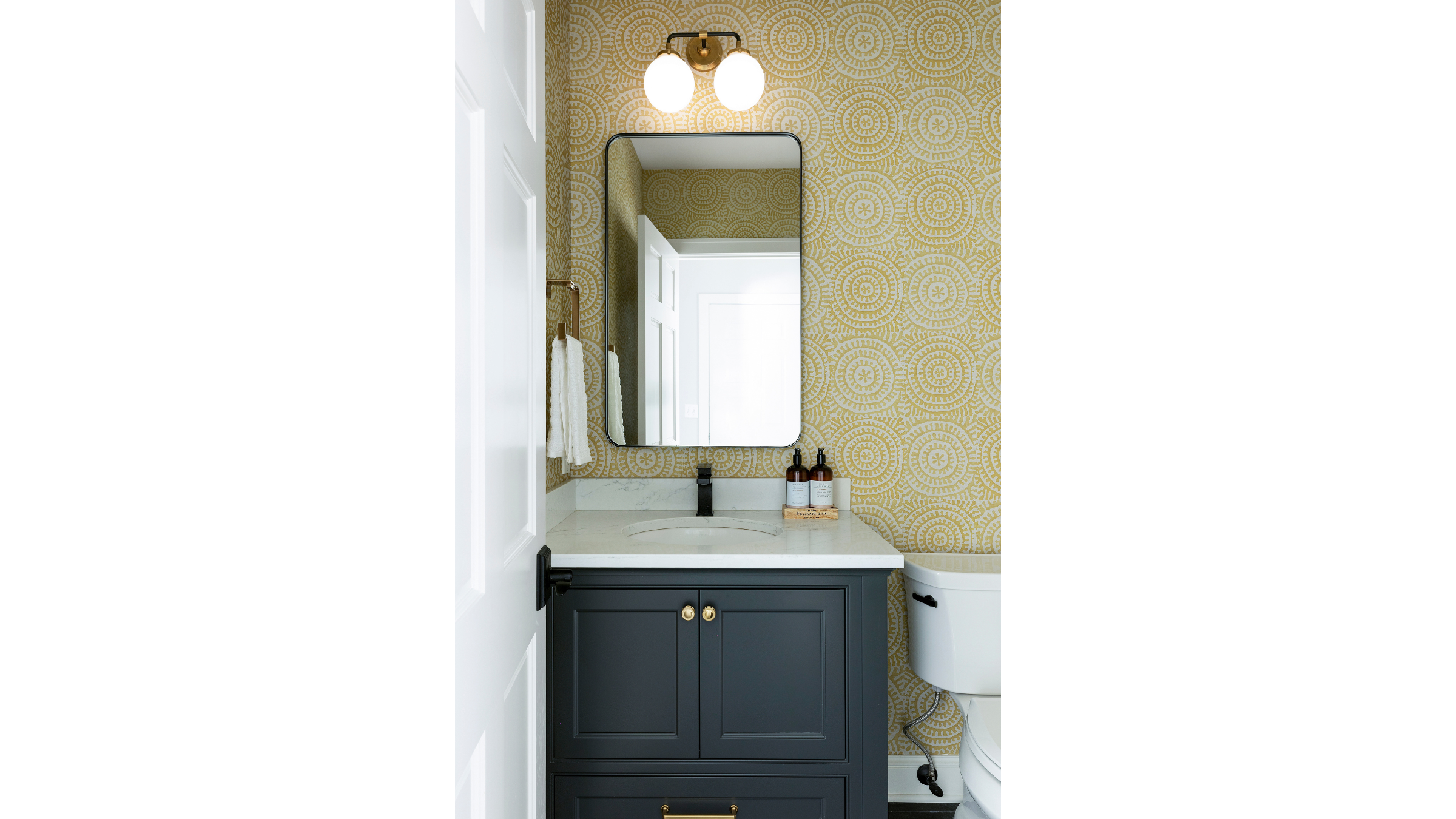 Bathroom vanity with dark blue cabinet, white marble countertop, and oval sink, with a large mirror, yellow patterned wallpaper, two amber glass bottles, towel, and toilet