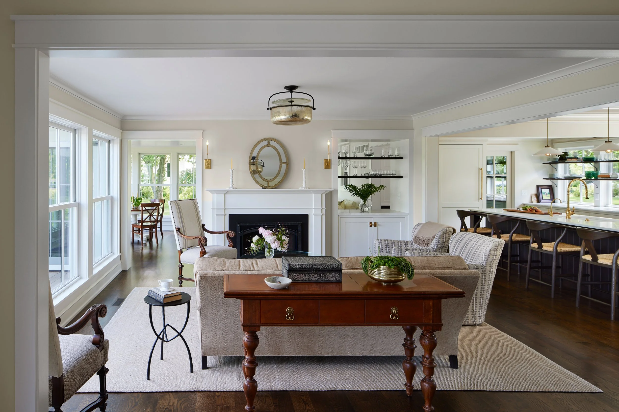 Living room with beige sofa, armchairs, a fireplace with mirror, and a dining area in the background, decorated in neutral tones with large windows and hardwood flooring.