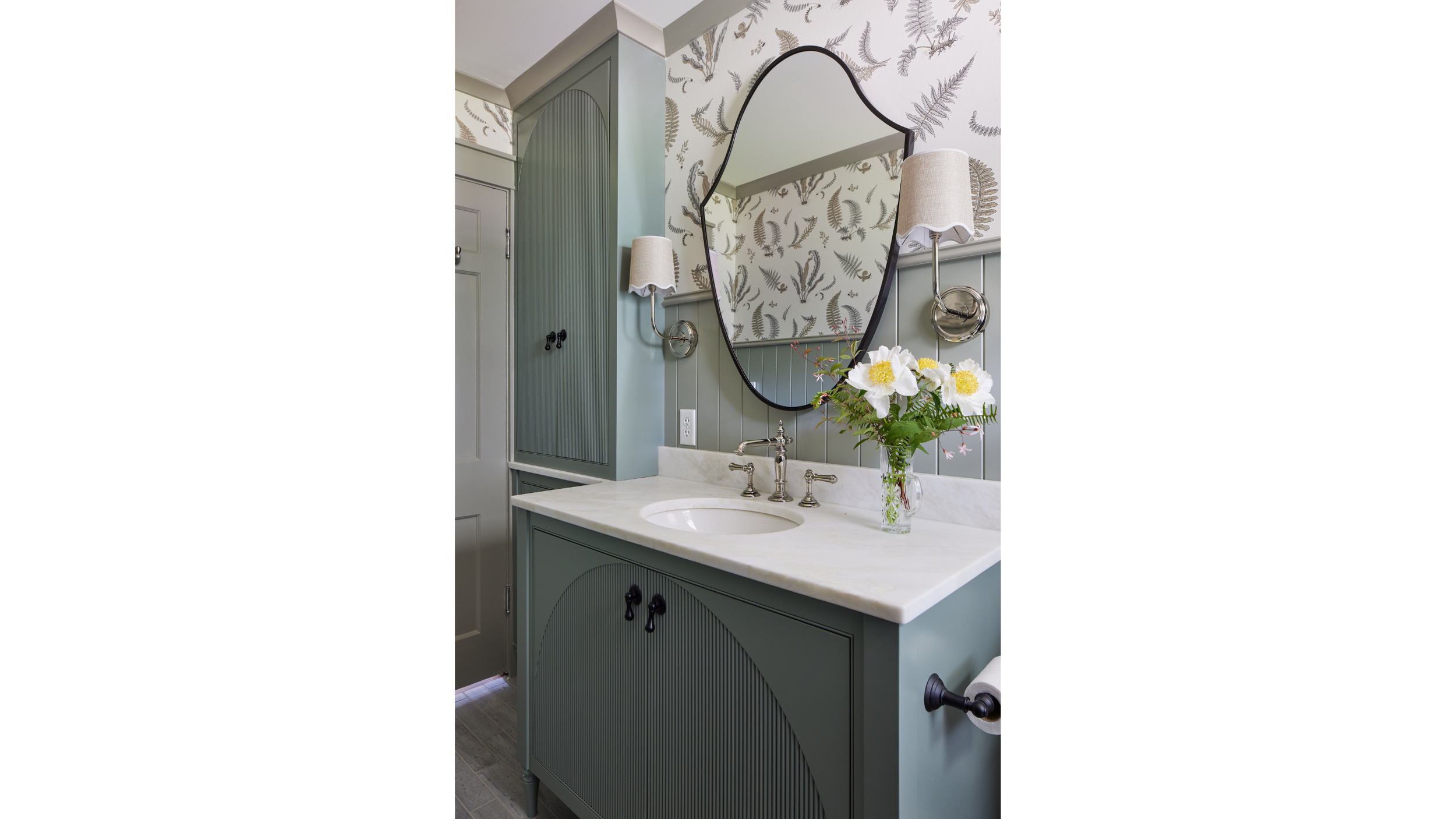 Bathroom with green vanity, marble countertop, oval mirror, and wall-mounted lamps, decorated with a vase of white flowers on the countertop.