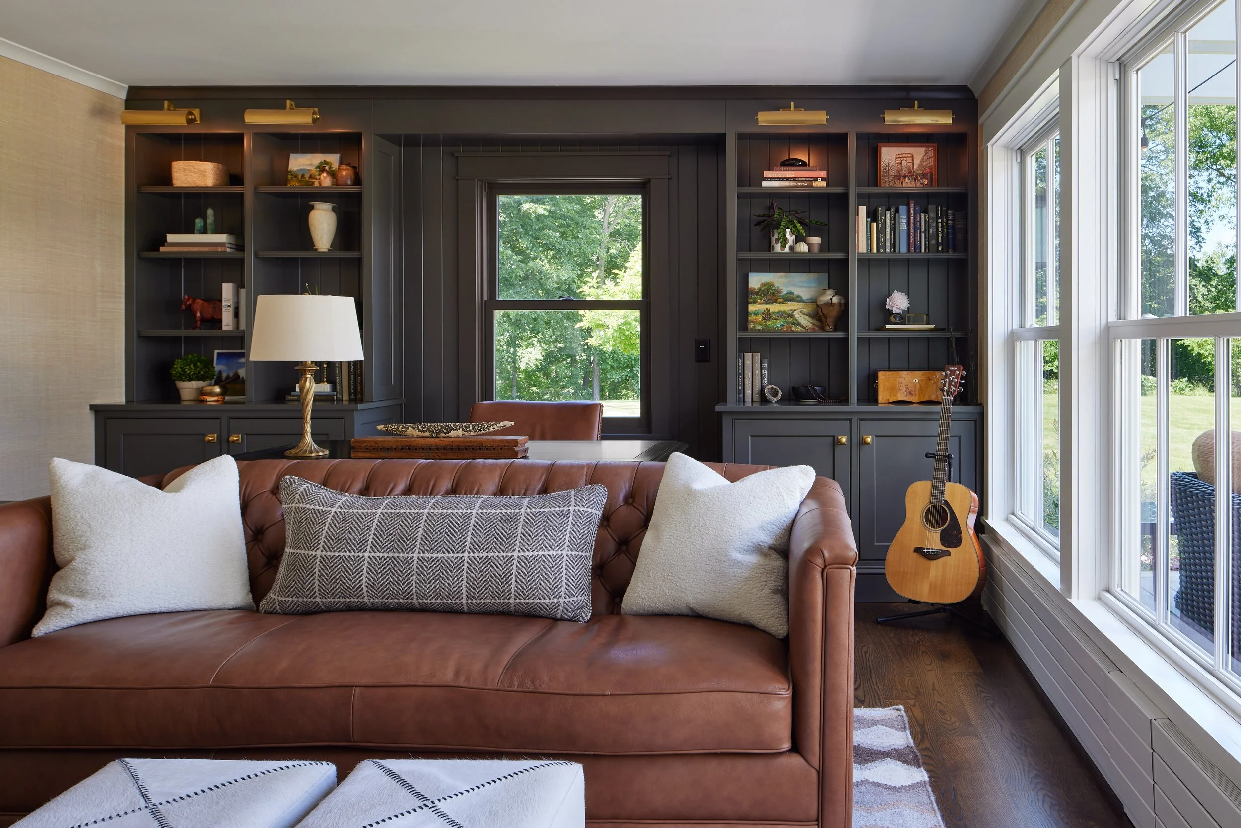 Living room with a brown leather sofa, white pillows, dark built-in bookshelves, a window with greenery outside, and an acoustic guitar near the window.