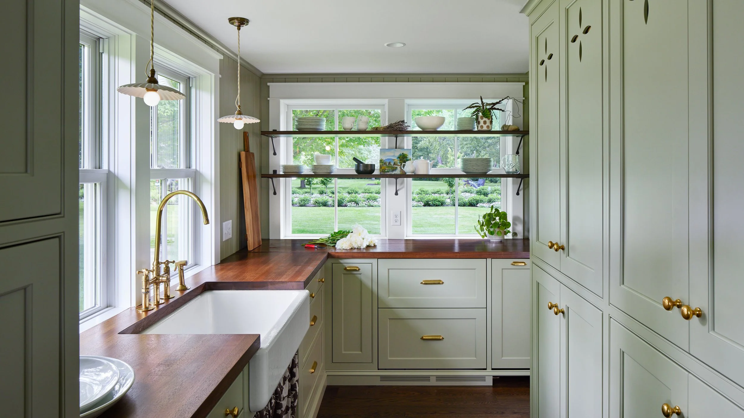 A cozy kitchen with light green cabinets, brass hardware, wooden countertops, a farmhouse sink, and a large window overlooking a green lawn with trees. Open shelves hold plates, bowls, and decorative items.