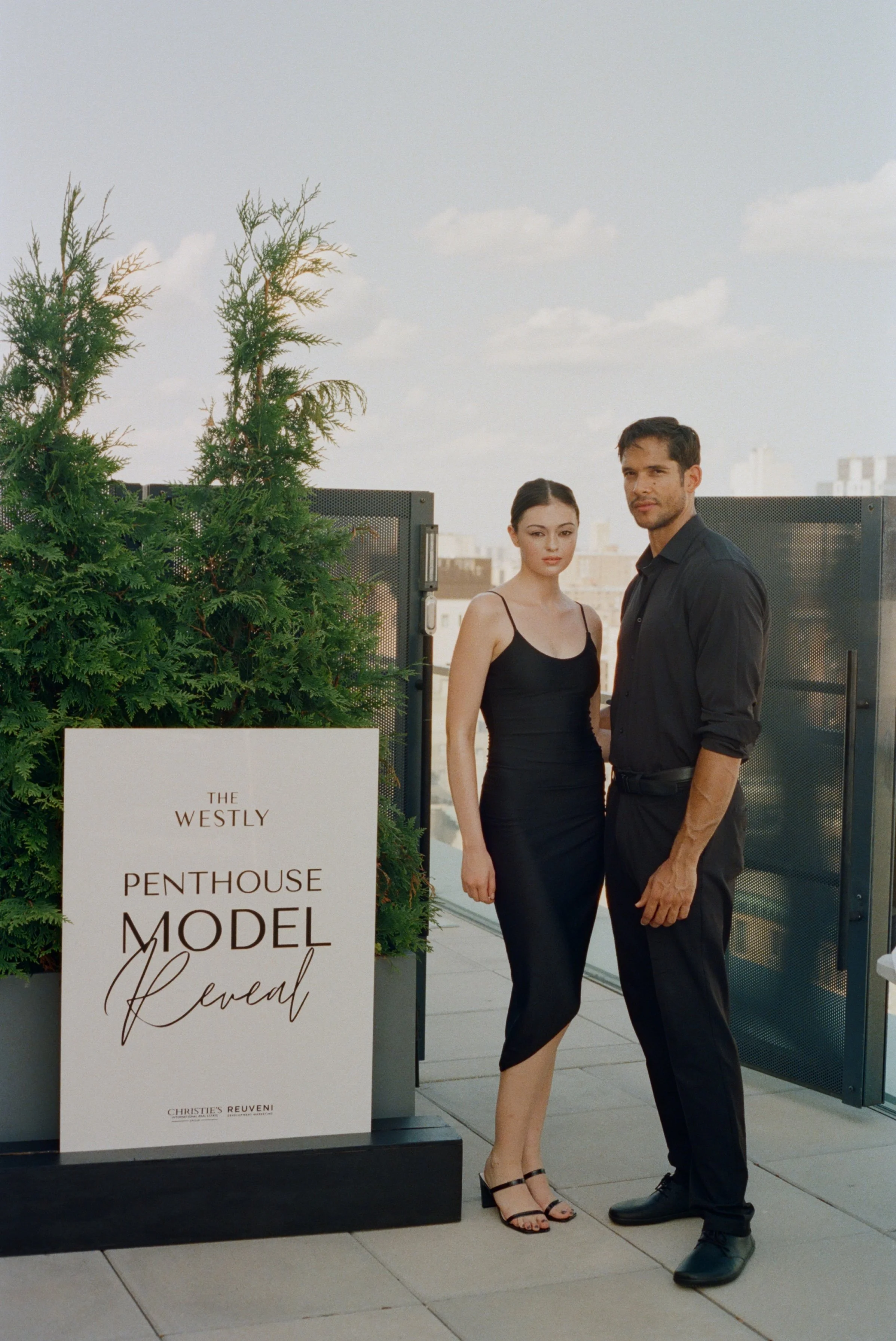 Una mujer y un hombre de pie en una terraza con un cartel que dice "The Westly Penthouse Model Lencial" y plantas verdes alrededor.