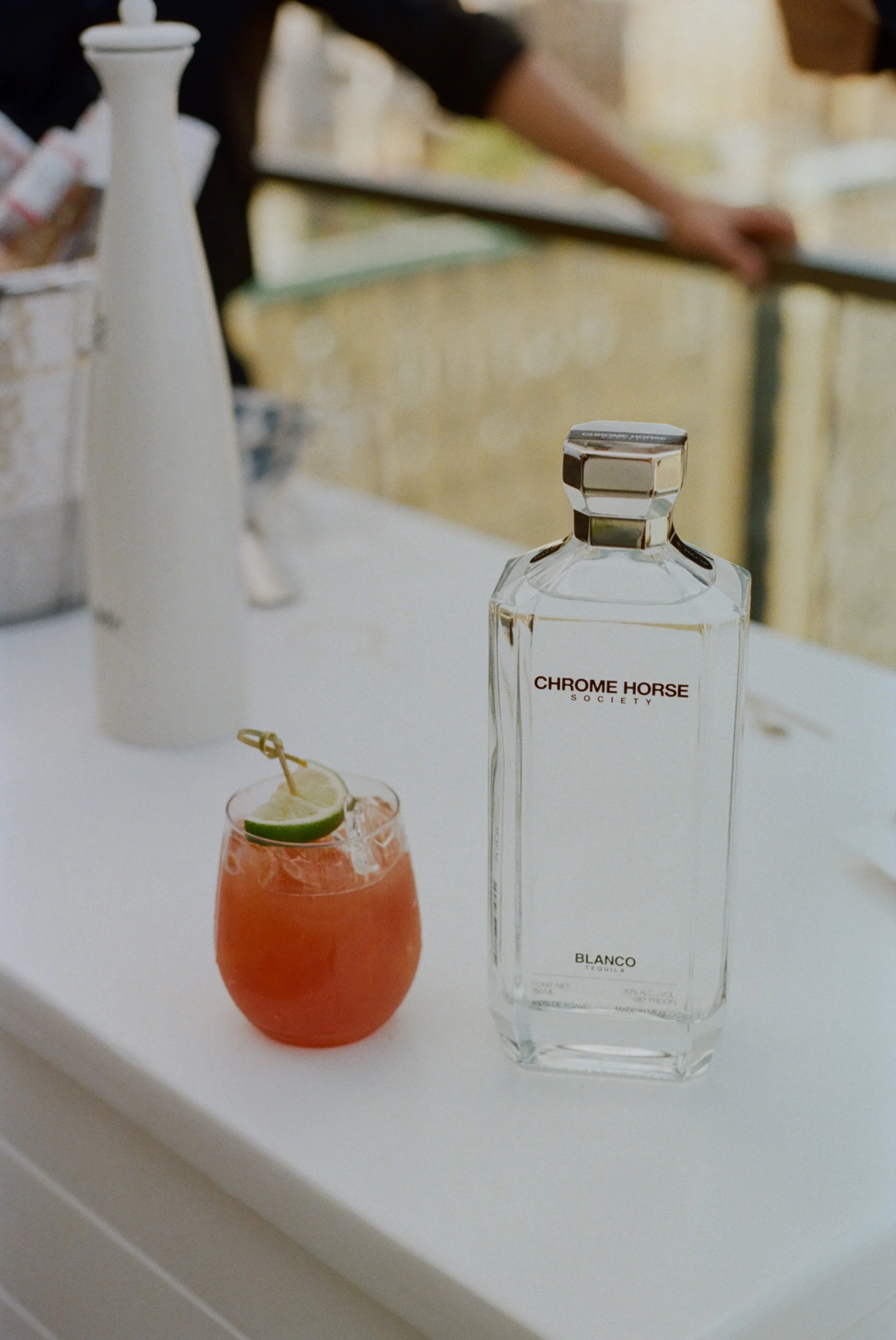 Una botella de tequila 'Chrome Horse Society Blanco' y un cóctel de color rojo con una rodaja de lima y una pajilla en una mesa blanca.