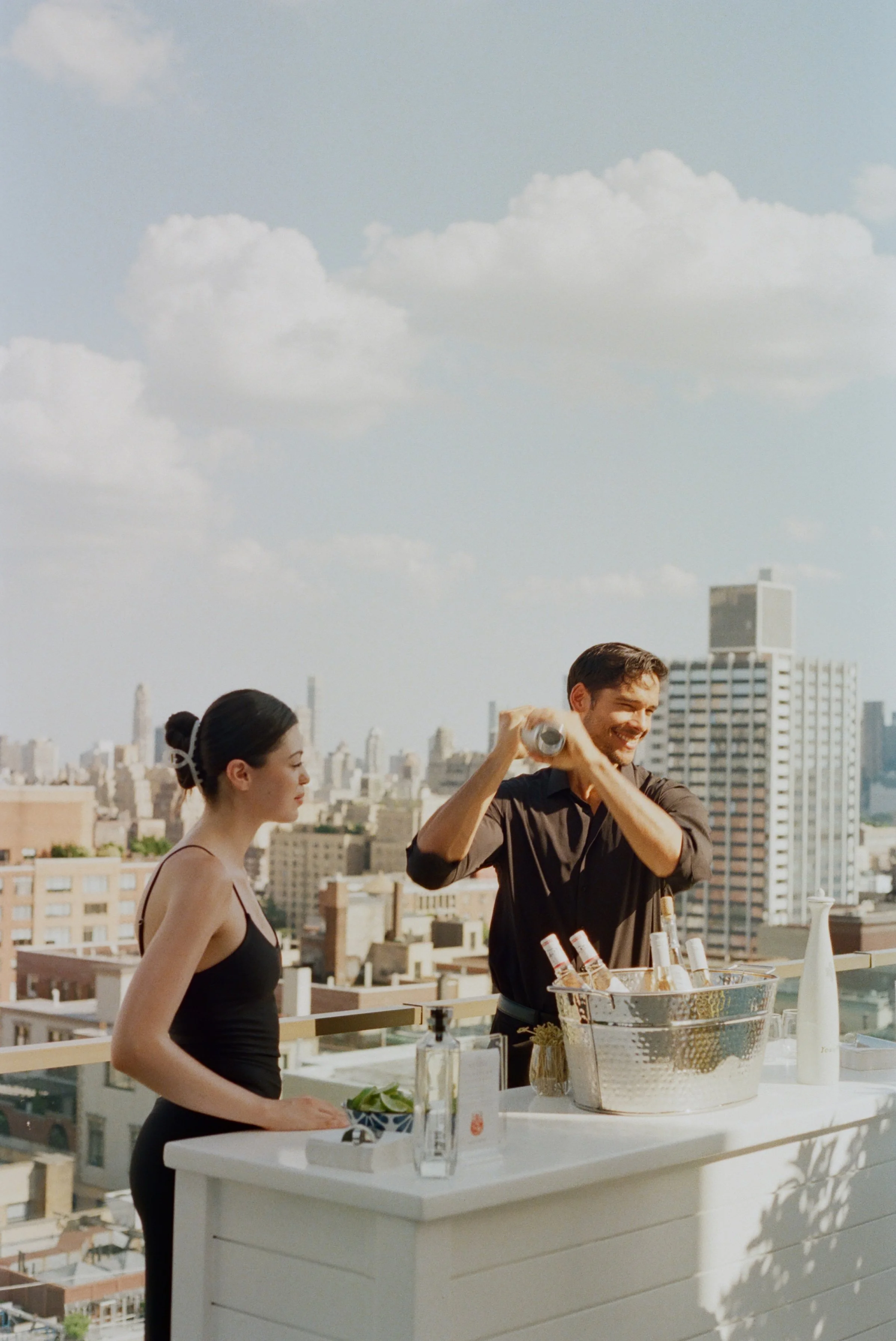 Un hombre y una mujer preparando bebidas en una terraza con vista a la ciudad en un día soleado.