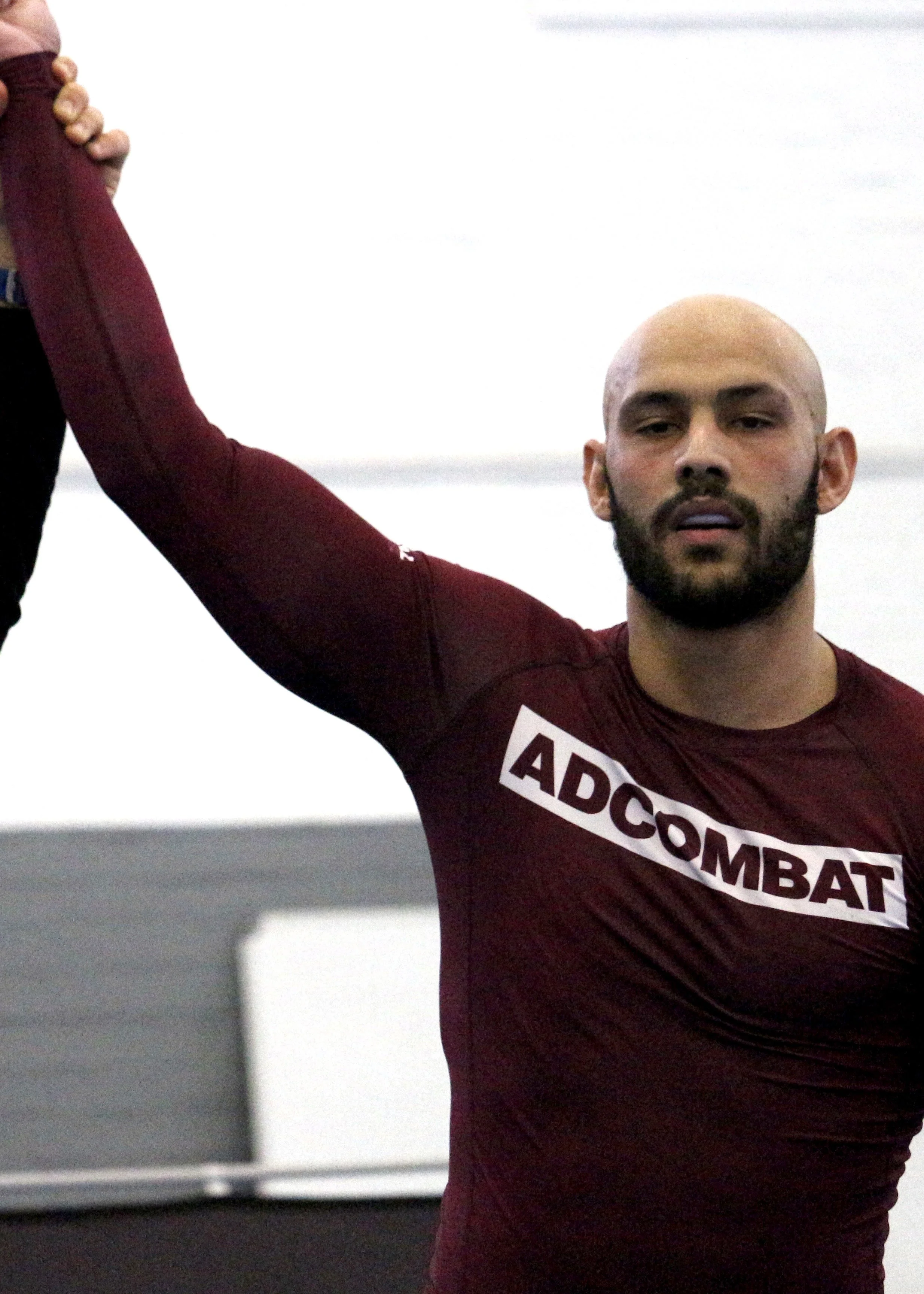 A man with a bald head and beard wearing a maroon athletic shirt with the word 'ADCOMBAT' on it, lifting his arm during a workout or training.