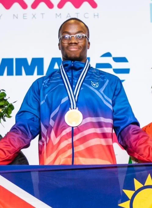 A man wearing glasses and a colorful sports jacket stands with a gold medal around his neck, smiling on a stage with MMA banners and a flag.