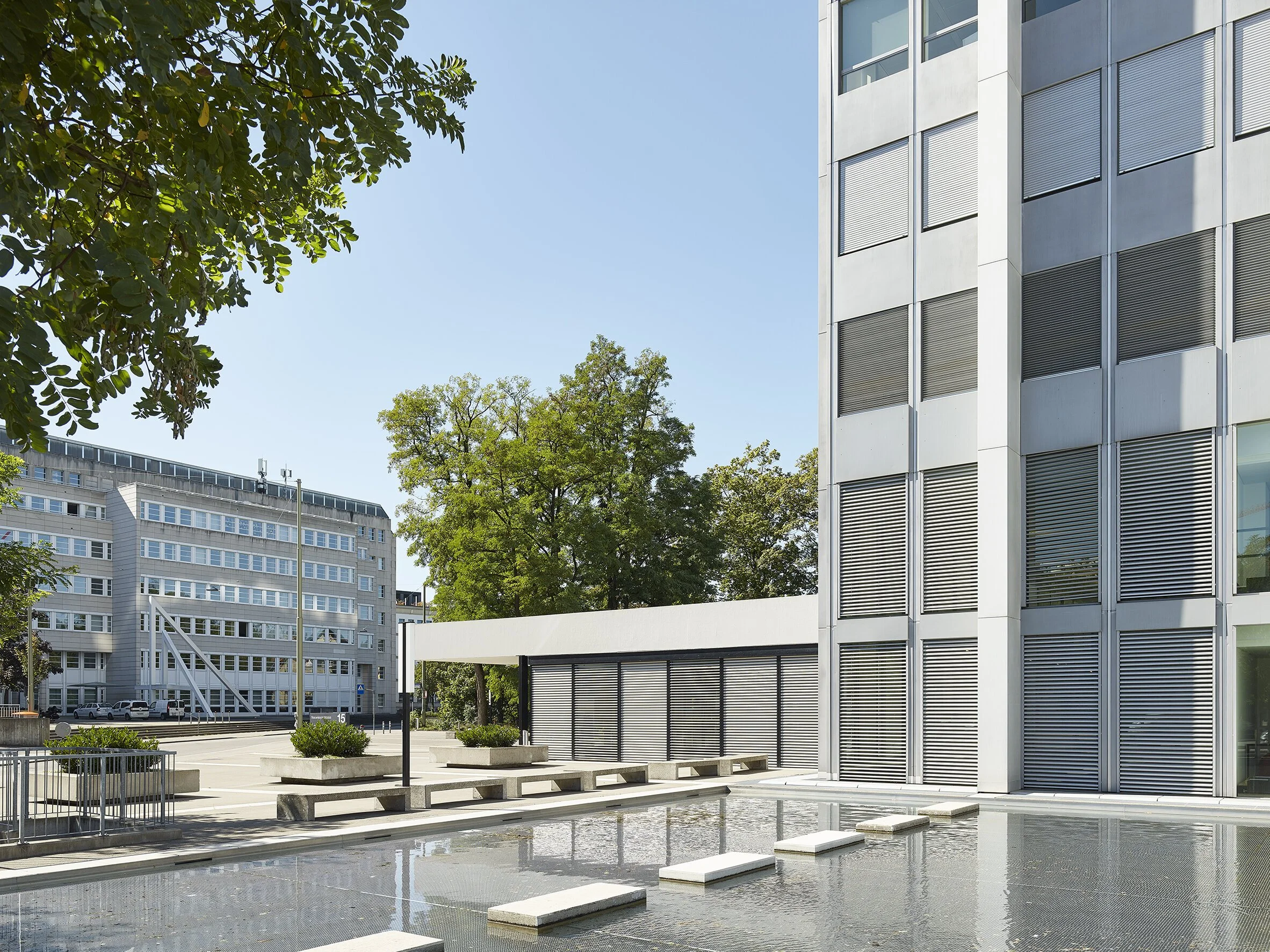 Moderne städtische Gebäude mit Wasserinstallation im Vordergrund, Bäume und blauer Himmel.