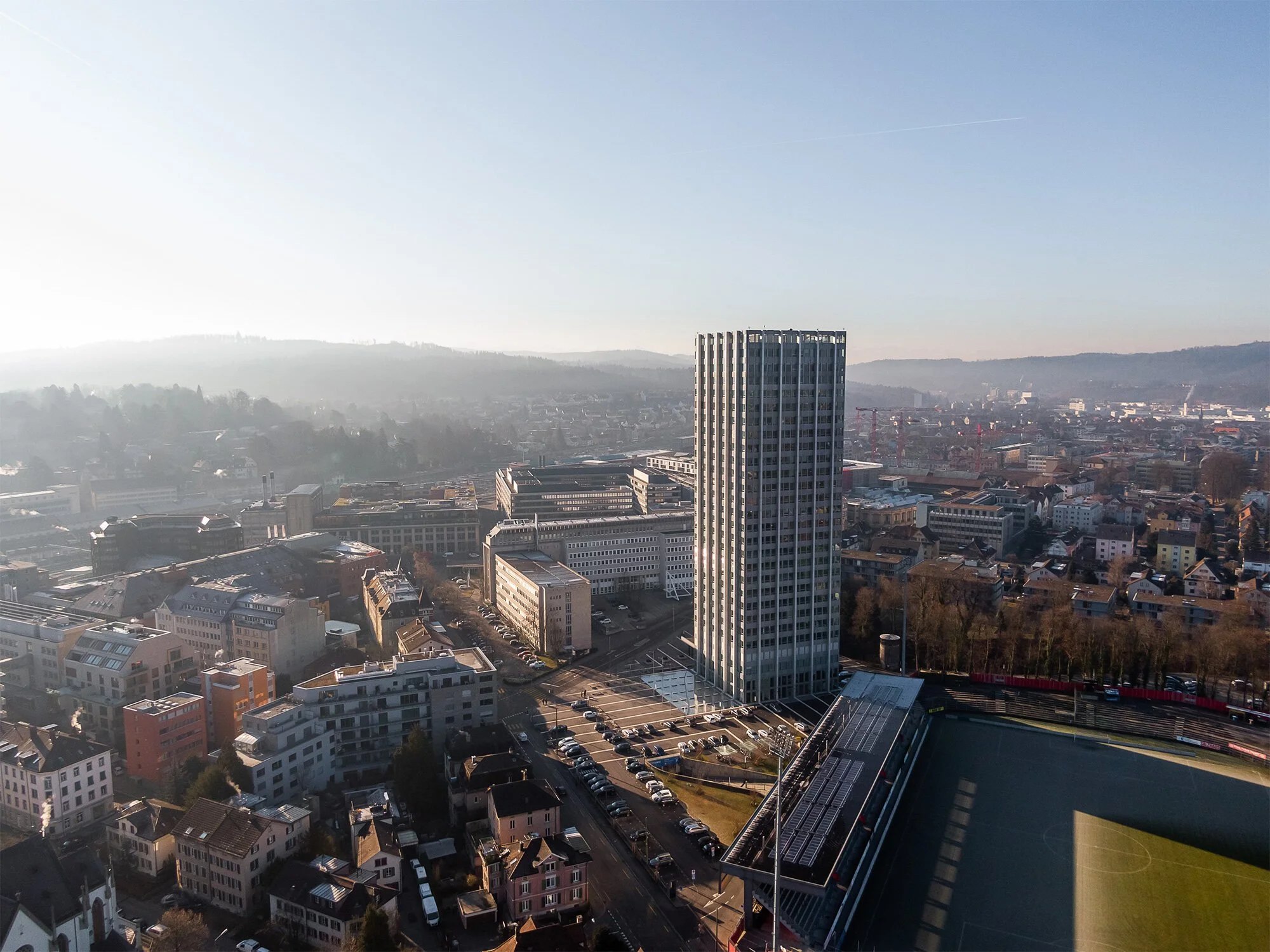 Stadtansicht mit hohem Bürogebäude, Wohnhäuser, Straßen und einem Fußballstadion bei Sonnenaufgang in einer europäischen Stadt.
