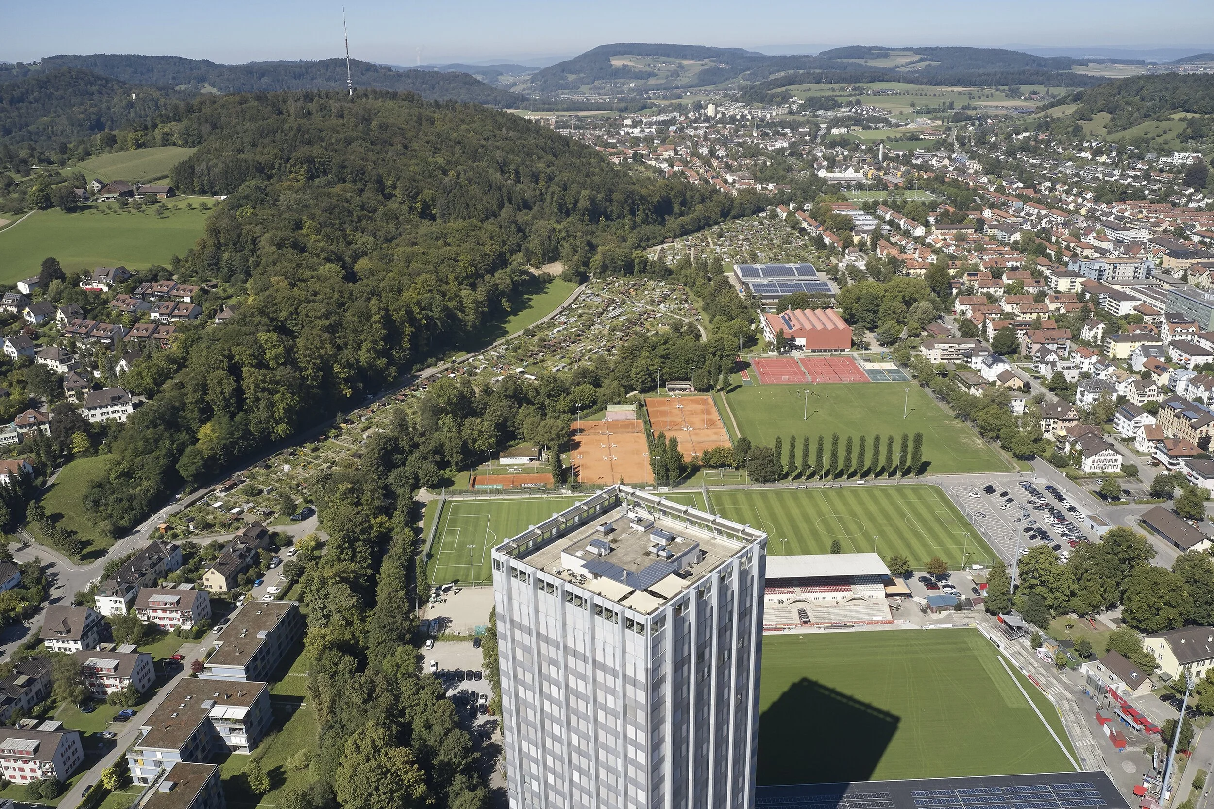 Luftaufnahme einer Stadt mit grünen Hügeln, Sportplätzen, Parks und einem Hochhaus.