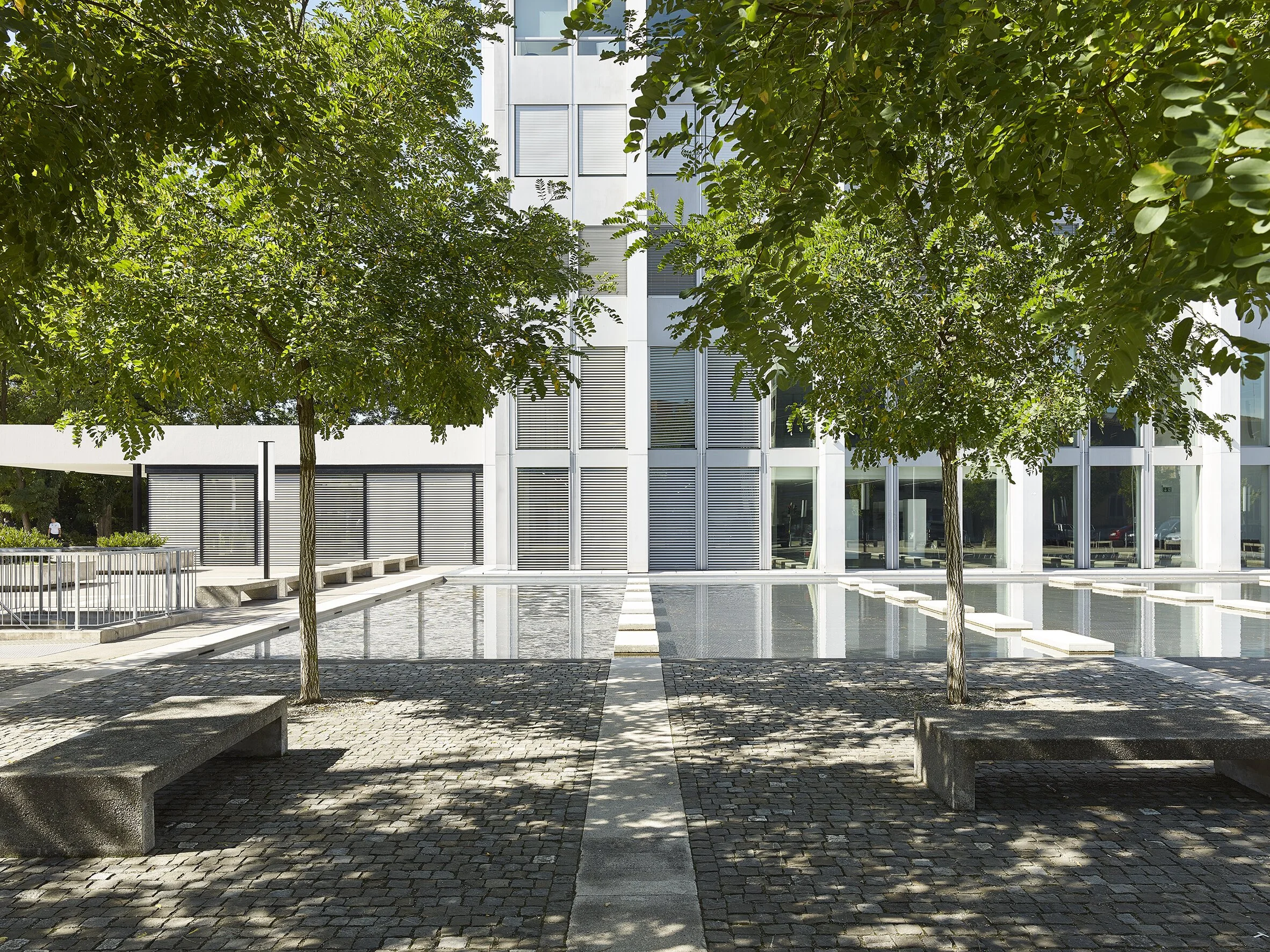 Moderne Gebäudefassade mit Glasfenstern, umgeben von Bäumen und Wasserbecken, mit Sitzbänken im Vordergrund.