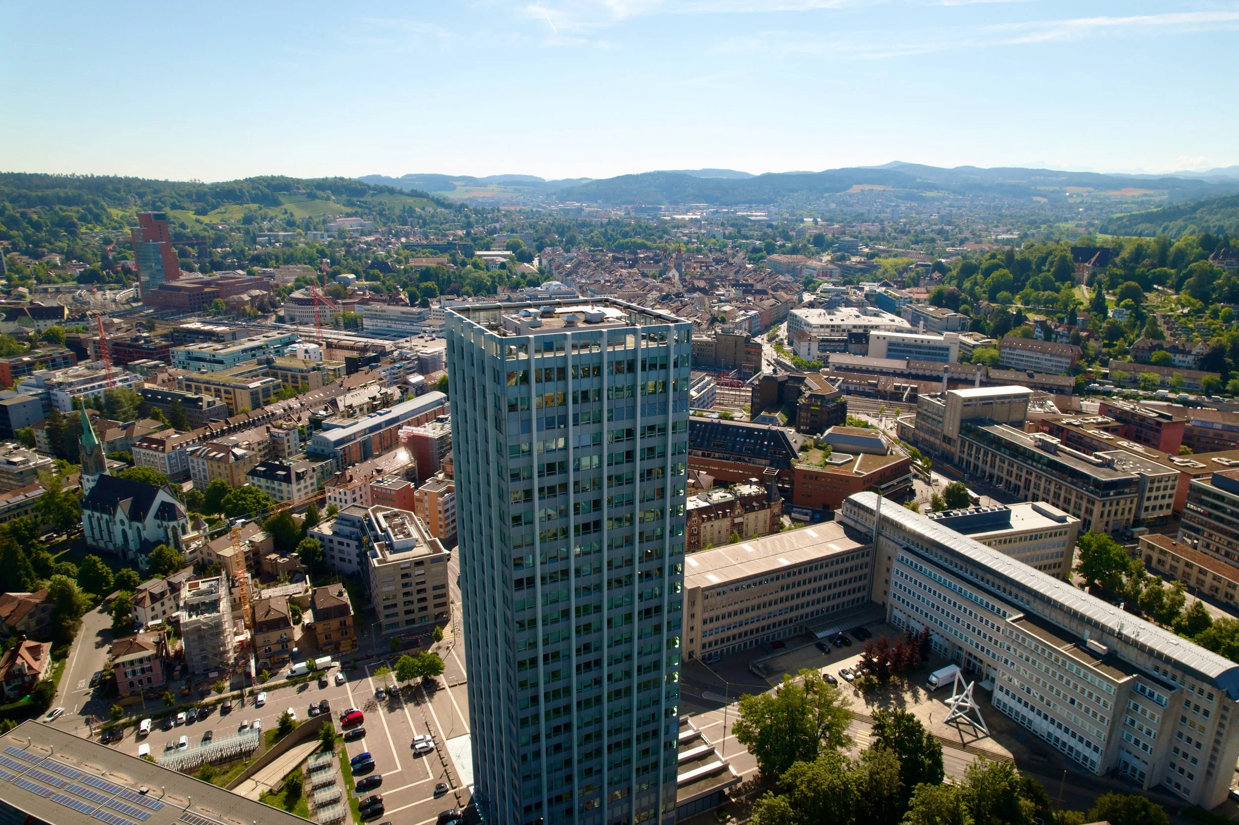 Stadtansicht mit hohen Gebäuden, einer Kirche mit grünem Turm, Parkplätzen und grünen Hängen im Hintergrund an einem klaren sonnigen Tag.