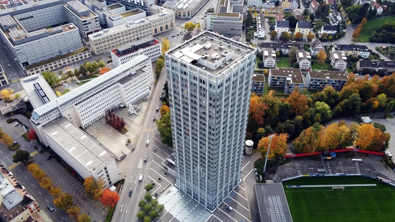 Ein hoher Wolkenkratzer in einer Stadt, umgeben von weiteren Gebäuden, Straßen, Bäumen mit Herbstlaub und einem Fußballstadion.