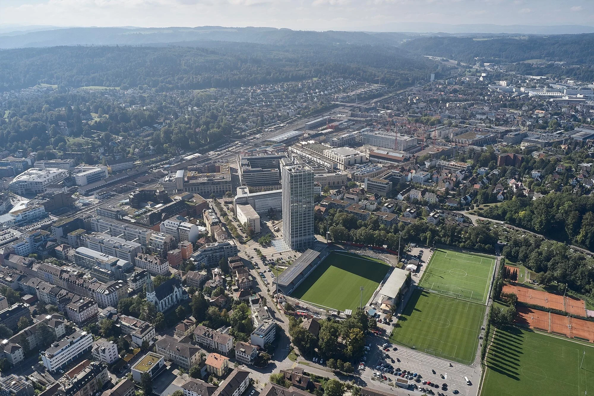 Luftaufnahme einer Stadt mit Gebäuden, Straßen, einem großen sportplatz mit Fußballfeldern und Tennisplätzen, umgeben von grünem Wald und Hügeln im Hintergrund.