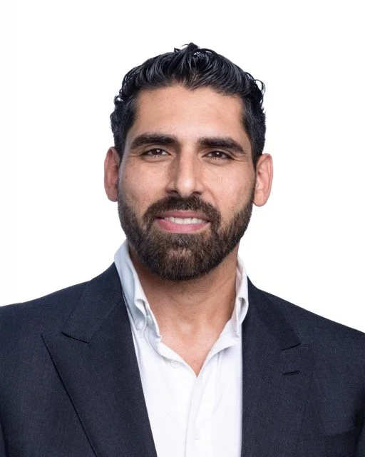 Close-up of a man with dark hair and beard, wearing a white shirt and maroon tie, smiling against a dark background.
