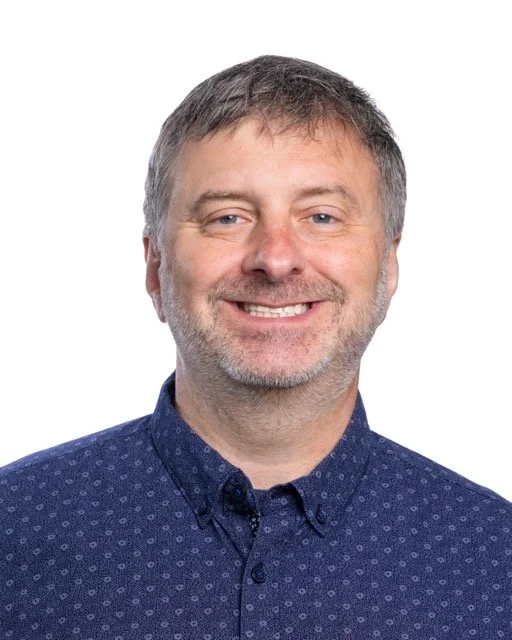 Smiling man with brown hair, light skin, wearing a white shirt and a tie, against a neutral background.
