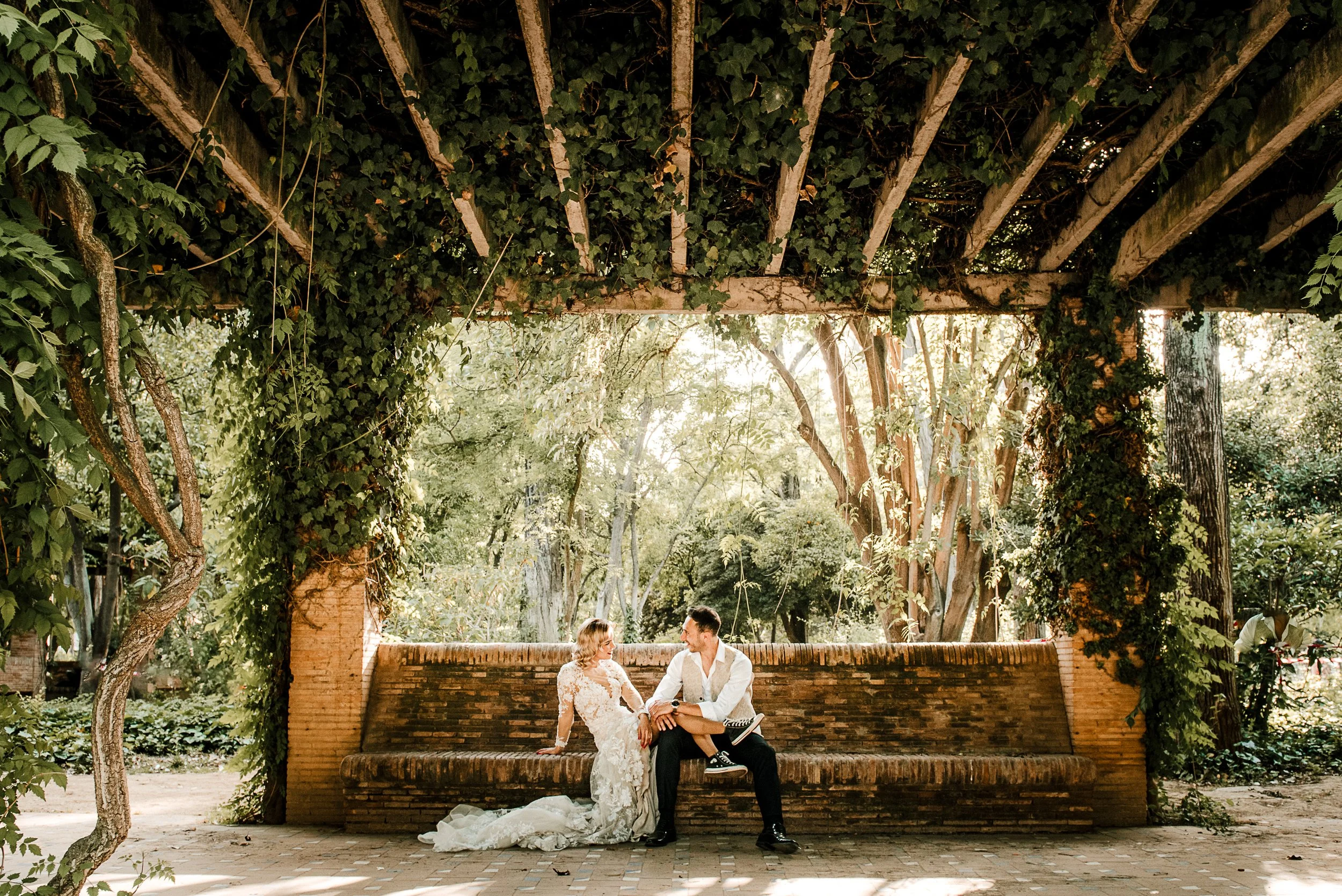 Pareja en vestido de boda y traje sentados en un banco de ladrillos bajo un dosel de madera cubierto de hiedra en un jardín con árboles.
