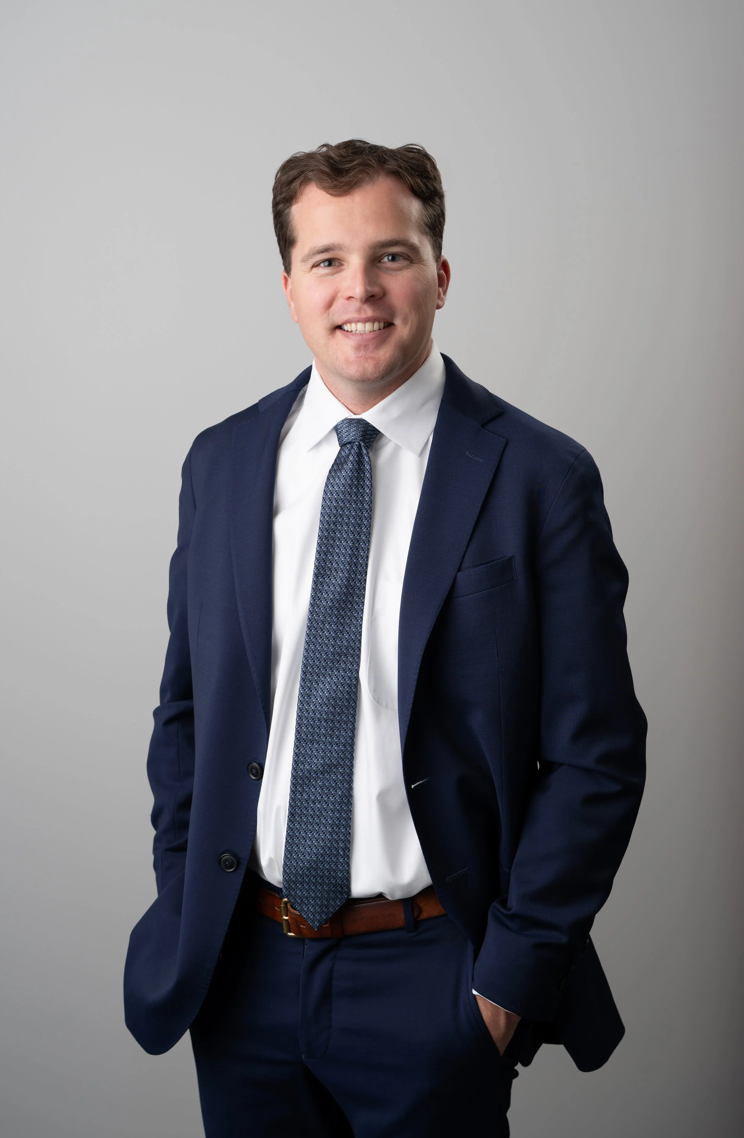 A young man in a navy blue suit, white shirt, and blue tie, smiling and posing with his left hand in his pocket against a plain gray background.