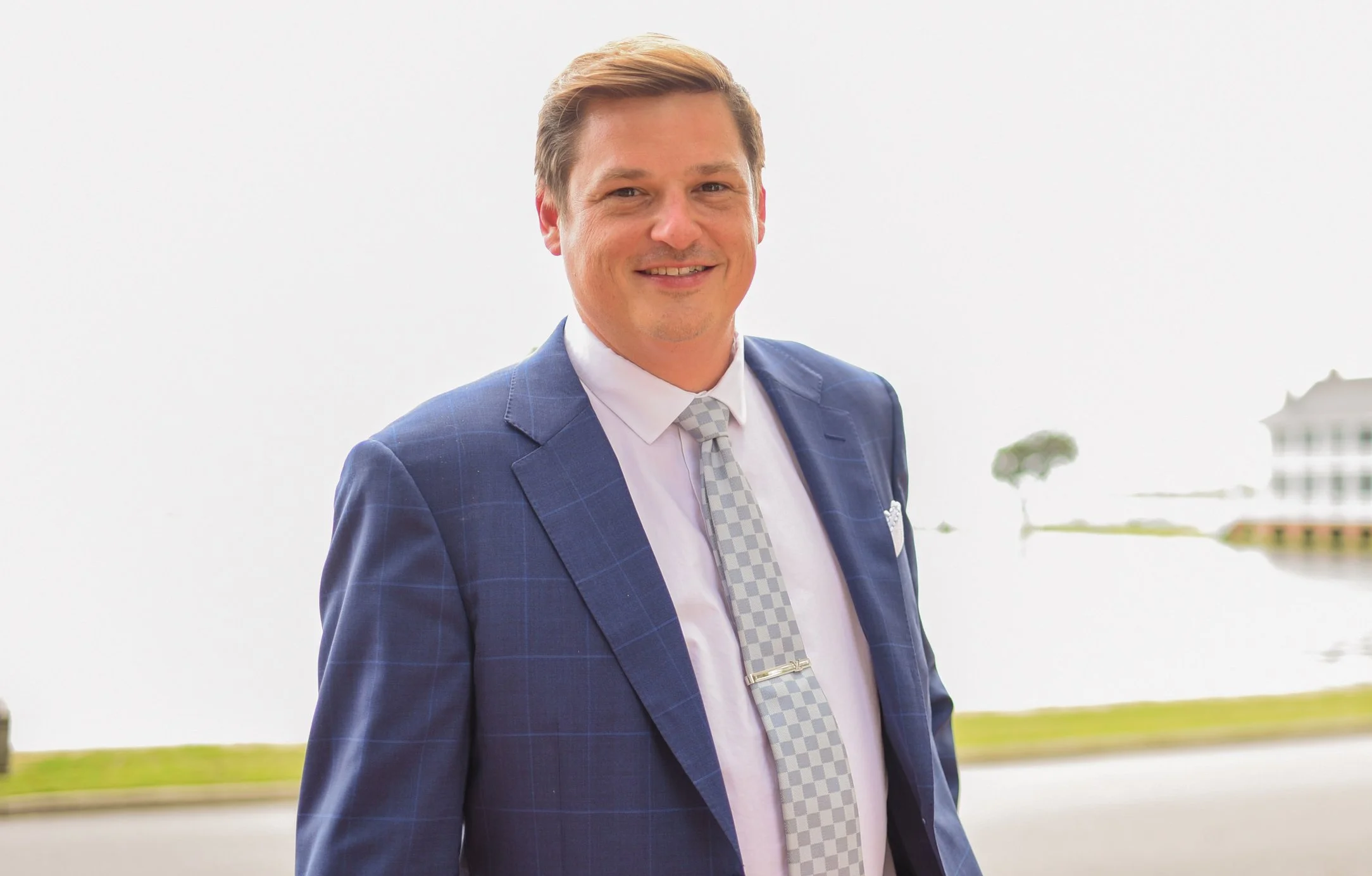 A man in a dark pinstripe suit, light blue shirt, and purple patterned tie, smiling outdoors in front of a modern sculpture, trees, flowers, and a house.
