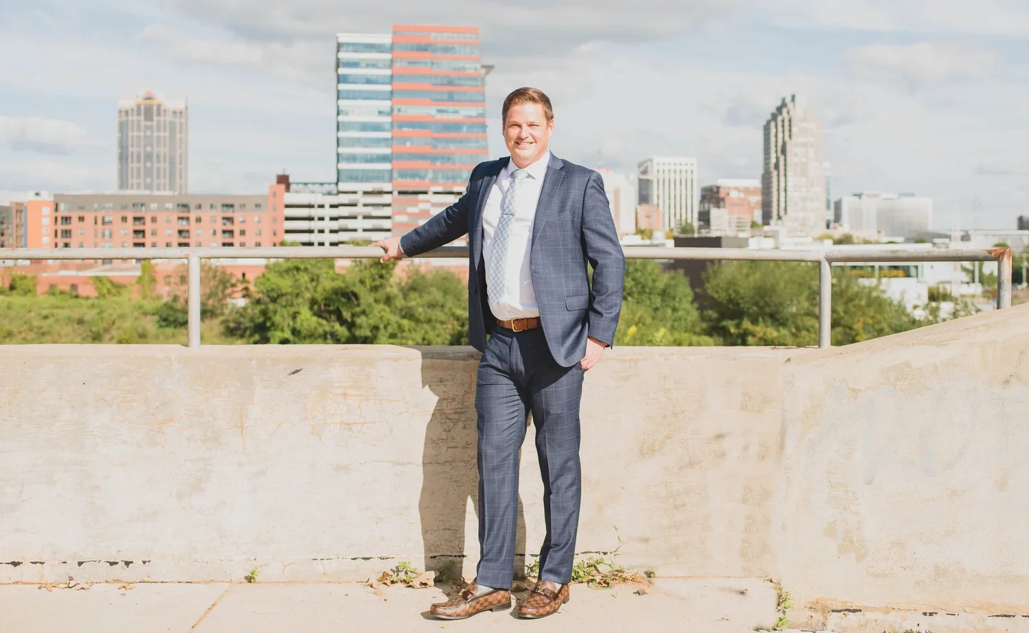 A man in a blue suit standing on a bridge with a city skyline in the background, smiling and resting on a metal railing.