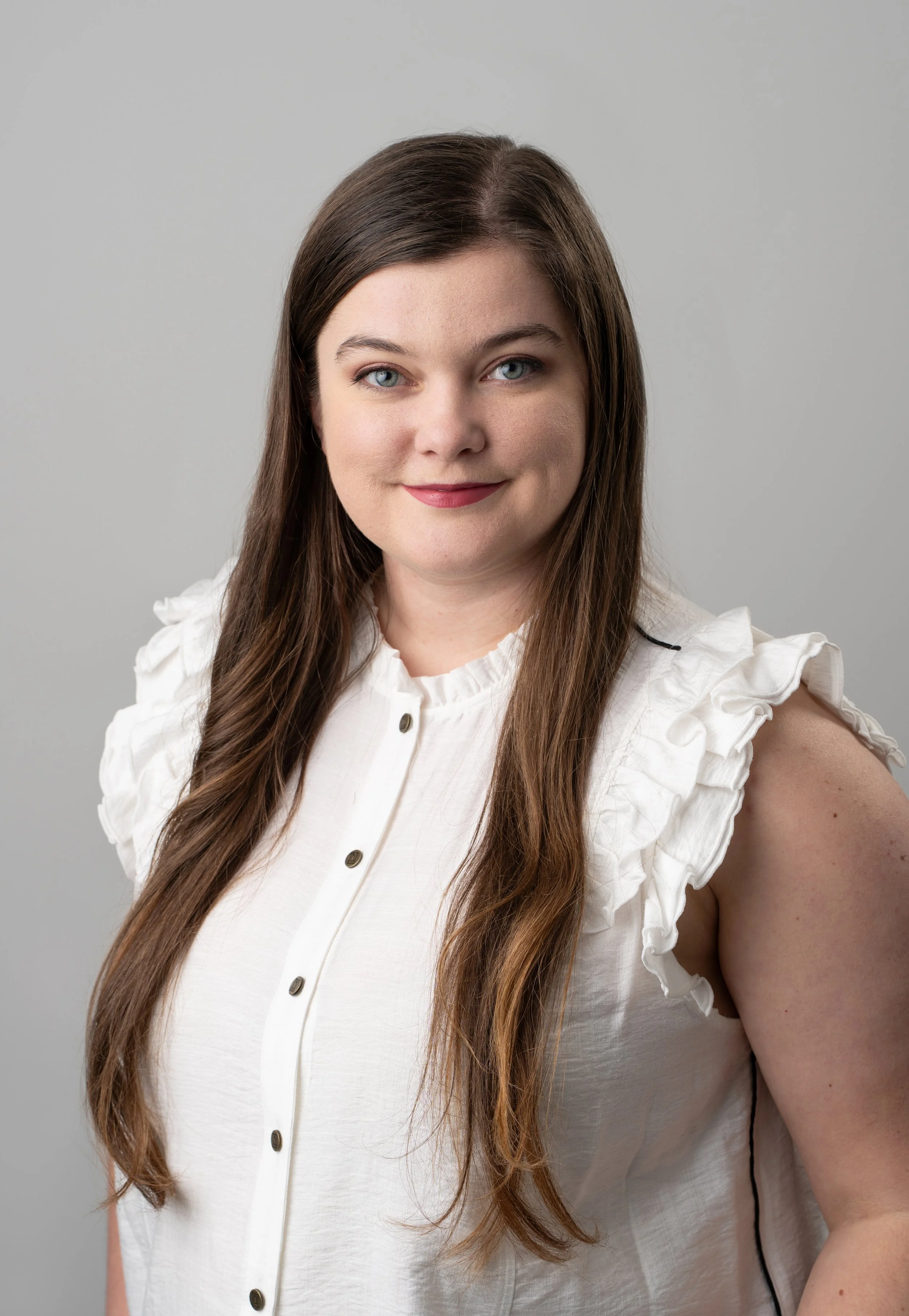 A young woman with long brown hair, blue eyes, and a slight smile, wearing a white sleeveless blouse with ruffled shoulders, standing against a plain gray background.