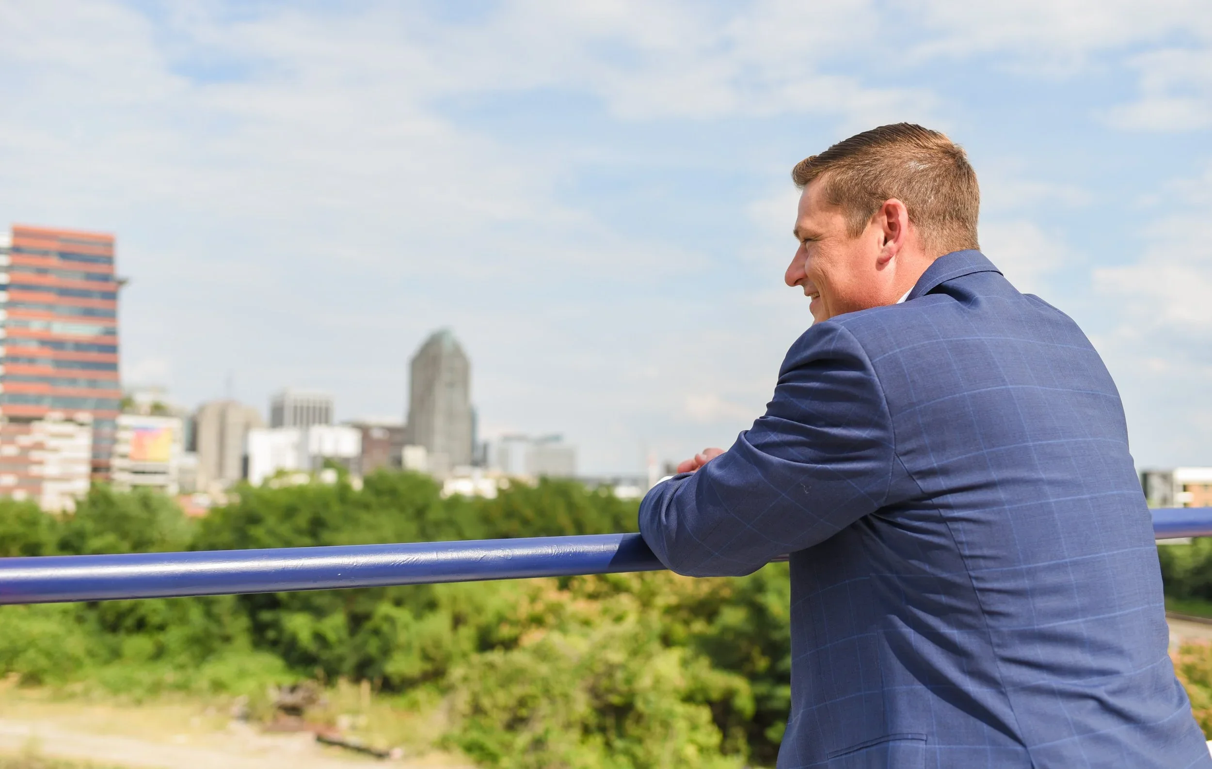 Man in a blue checkered suit leaning on a metal railing and smiling while looking out at the city skyline with trees in the foreground and a partly cloudy sky.