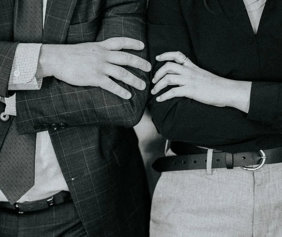 Close-up of two people with arms crossed, wearing business attire; one in a checked blazer and the other in a dark blouse.
