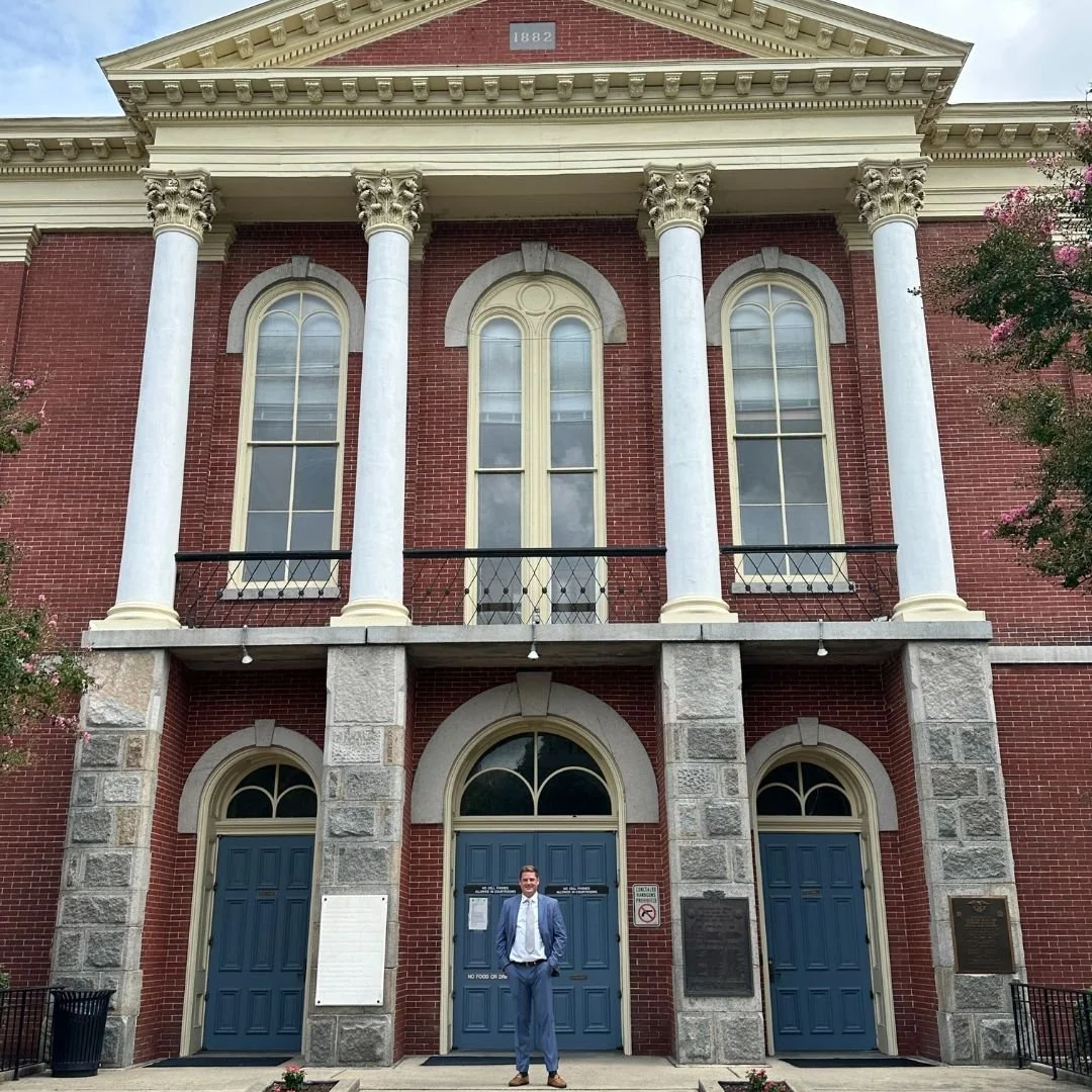 A man in a suit standing in front of a historic red brick building with large arched windows, tall white columns, and plaques on either side of the entrance, under a pediment with the year 1882.