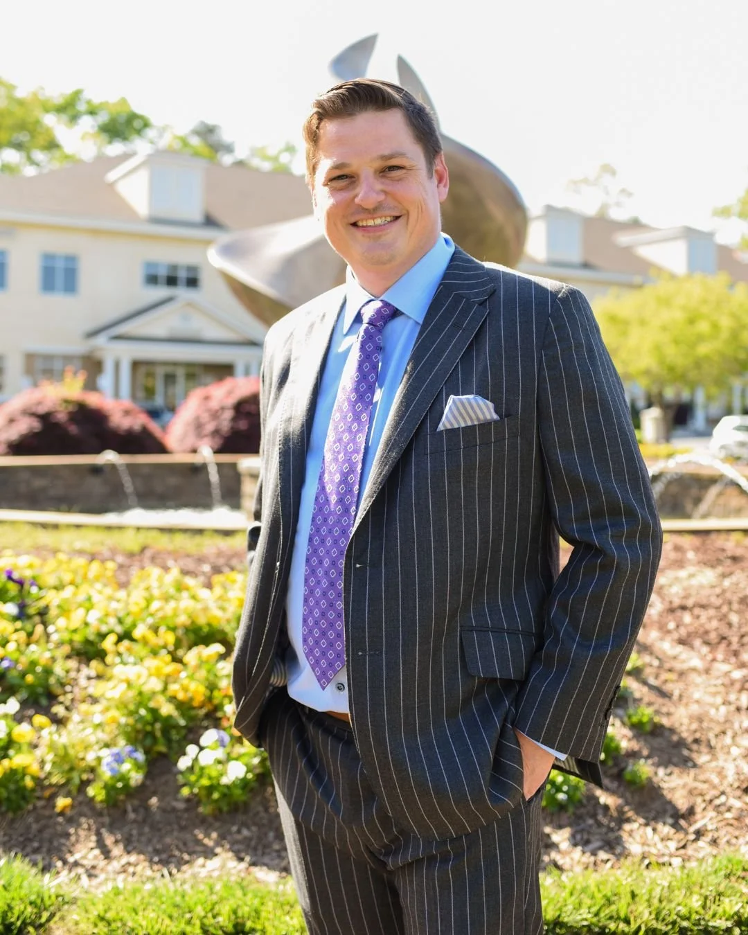A man in a dark pinstripe suit, light blue shirt, and purple patterned tie, smiling outdoors in front of a modern sculpture, trees, flowers, and a house.