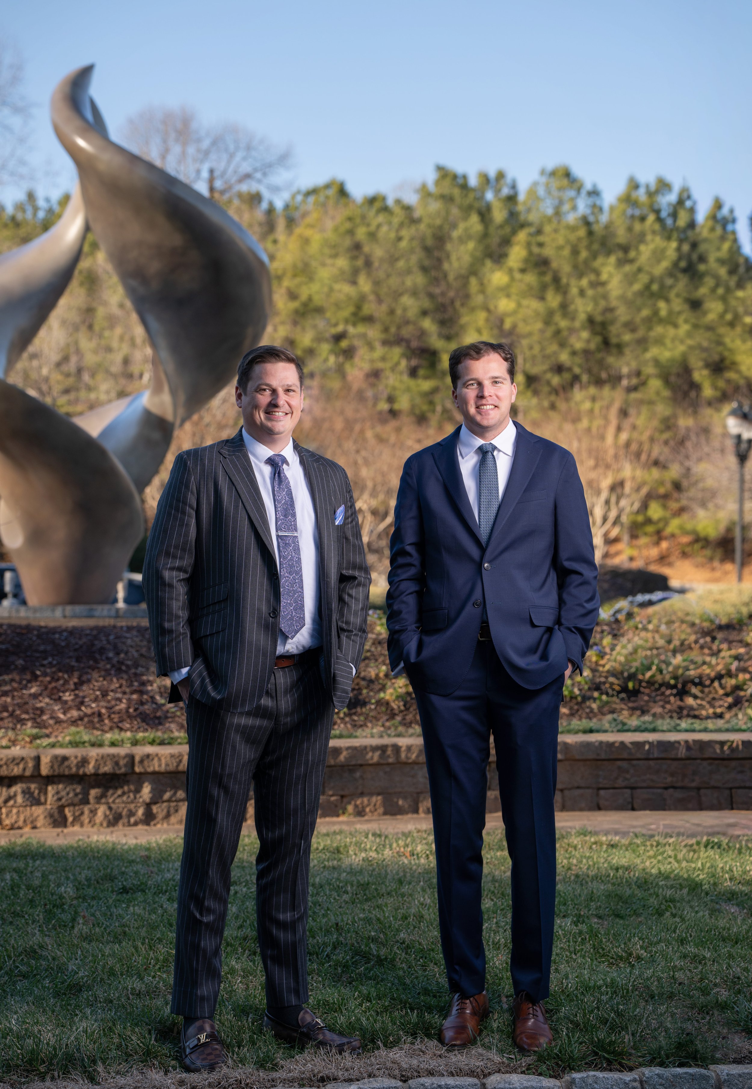 Two men in suits standing outdoors in front of a sculpture and trees, smiling at the camera.