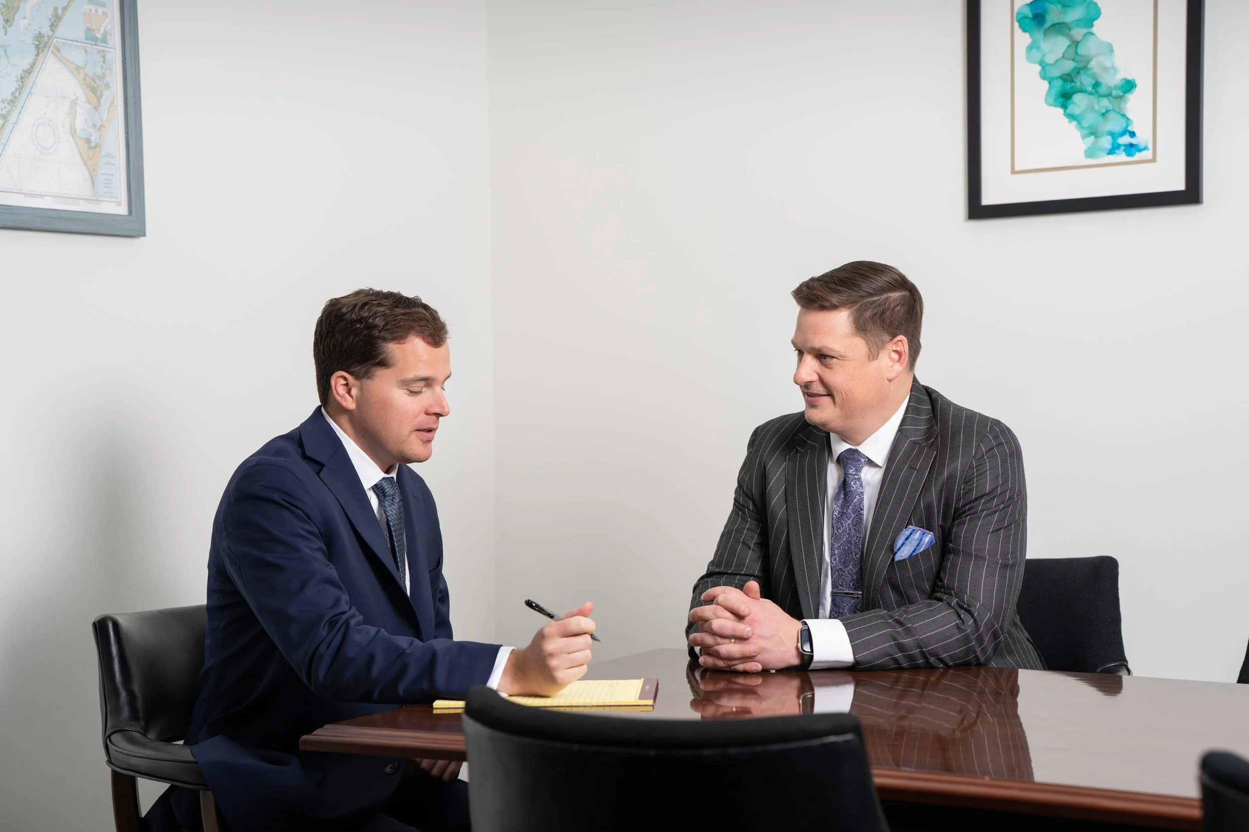 Two businessmen sitting across from each other at a conference table having a discussion, one holding a pen and the other with hands clasped, in a professional office setting.