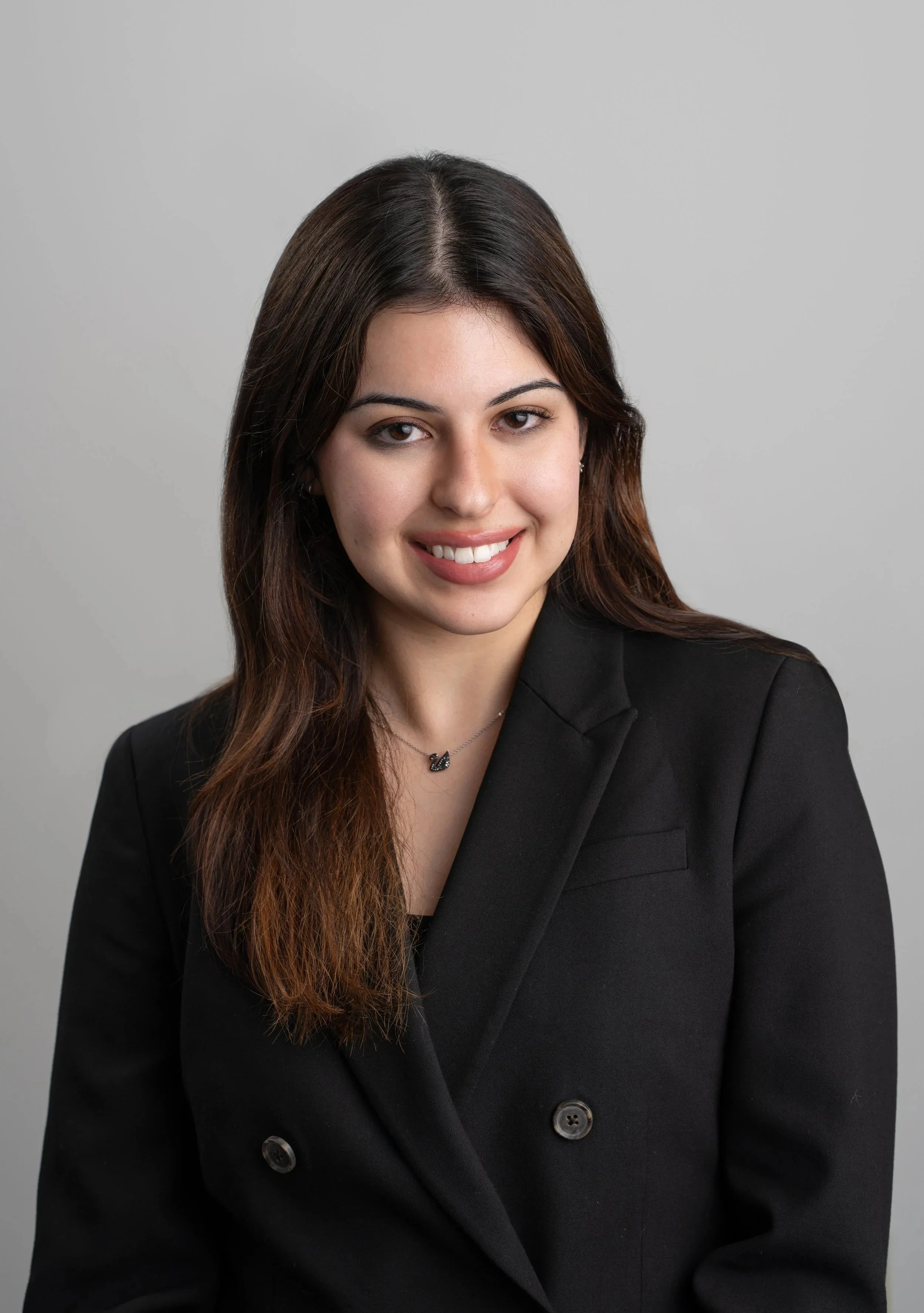 Professional portrait of a young woman with long brown hair, smiling, wearing a black blazer and a necklace with a small pendant, against a plain gray background.