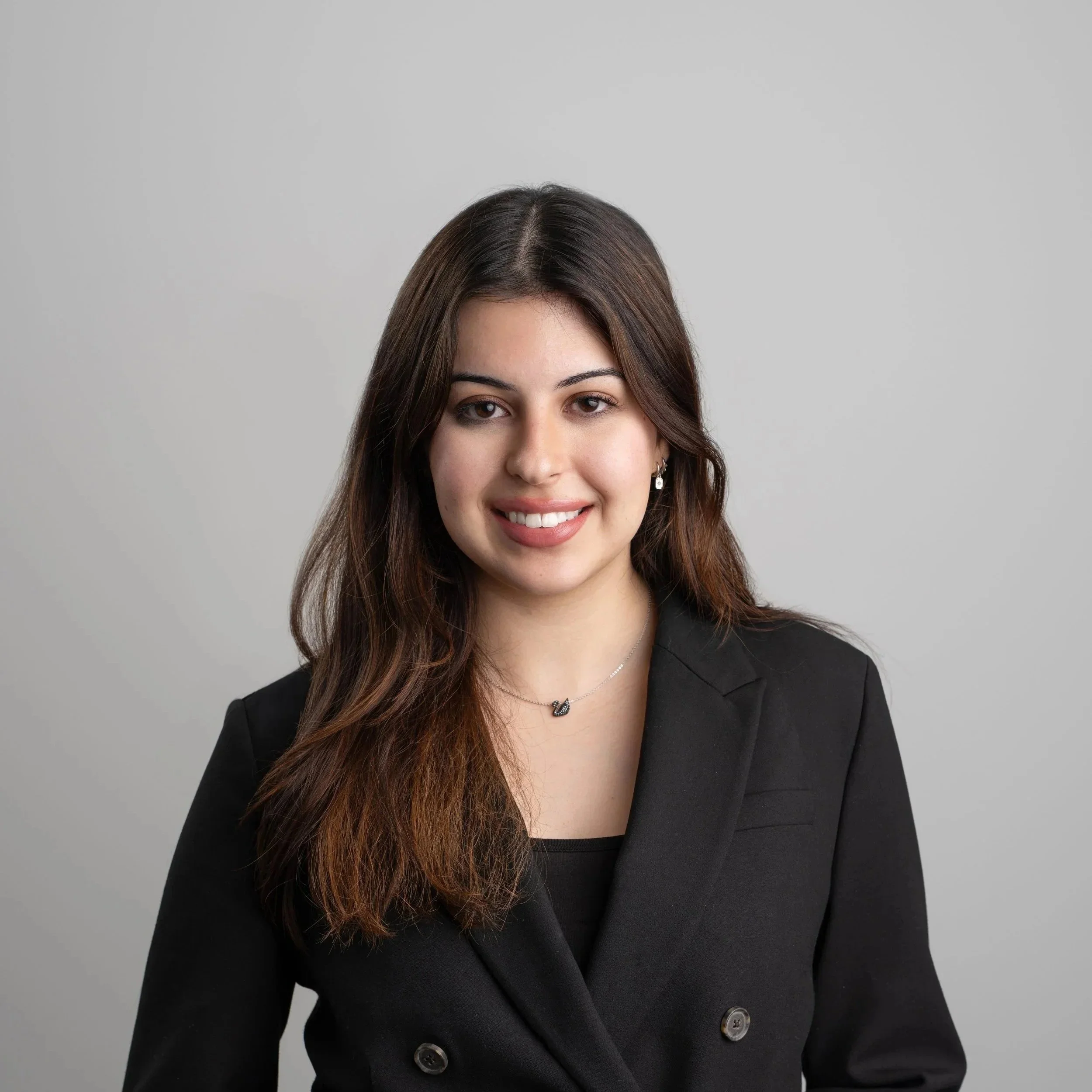 A young woman with long dark hair standing outdoors in a garden with colorful flowers, smiling, with houses and trees in the background.