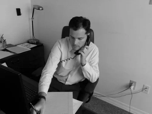 A man sitting at a desk talking on a landline phone, with a computer and papers in front of him, in a small office with a lamp and a side table.