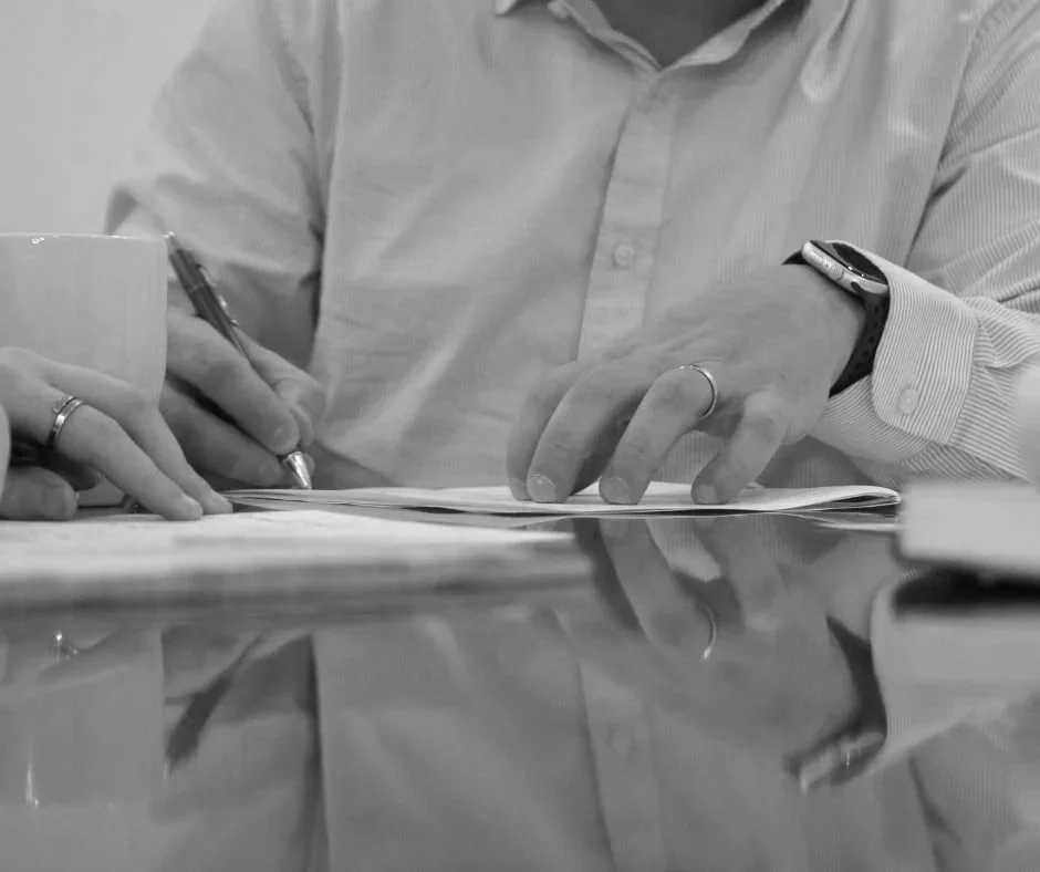 A person in a button-up shirt is sitting at a table, writing on paper while holding a pen in one hand. A cup and a watch are also visible on the table.