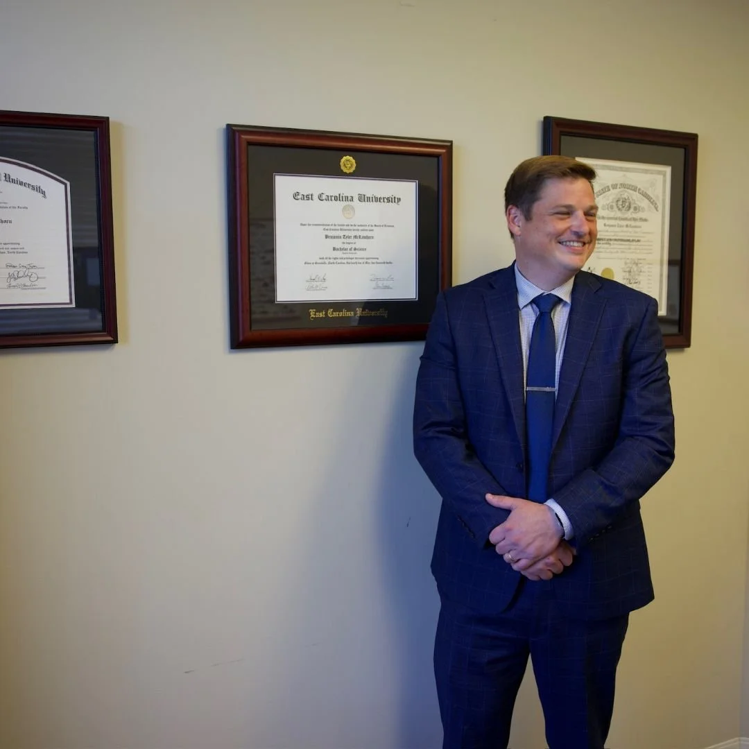A man in a blue suit and tie smiling, standing in front of a wall with framed diplomas and certificates.