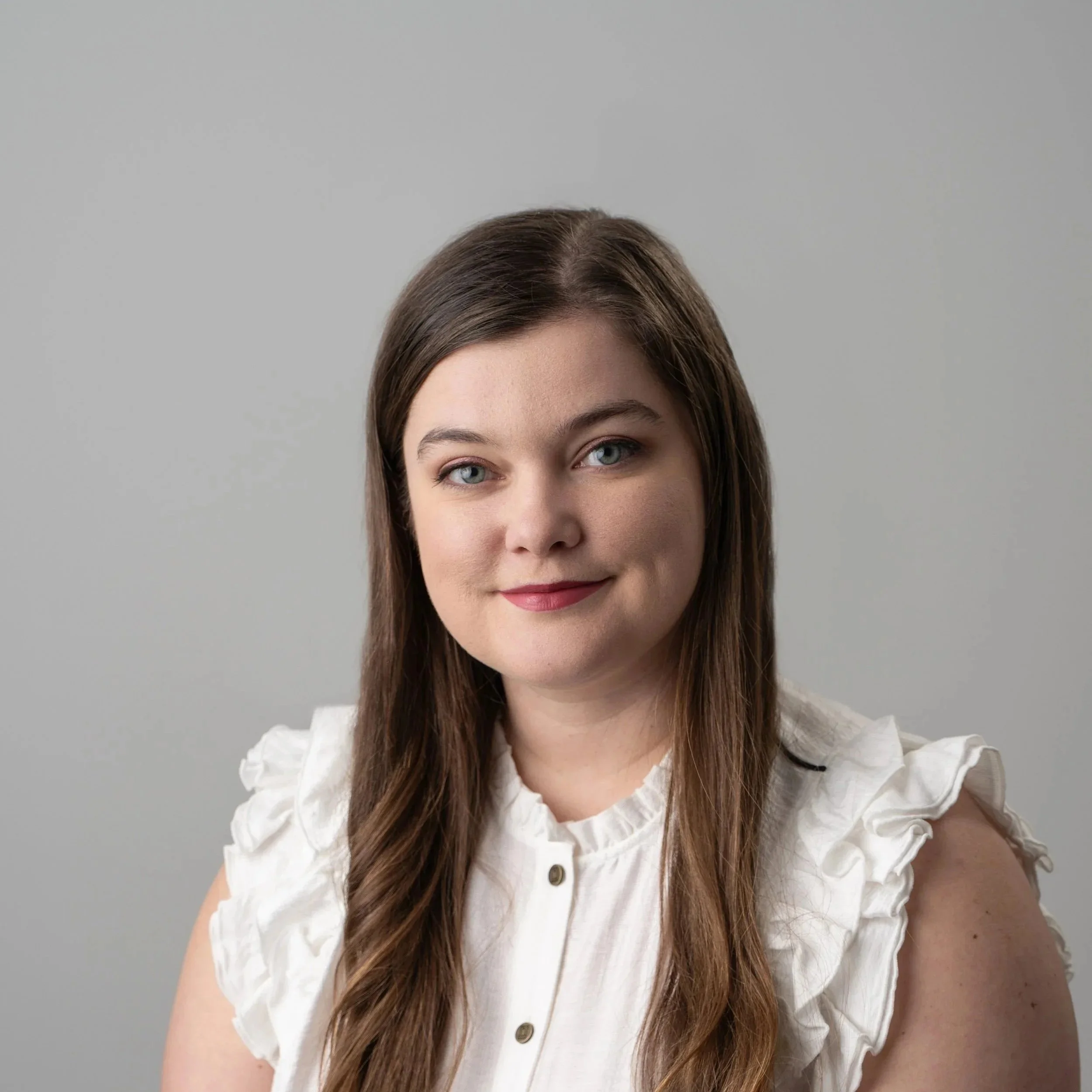 A woman with long brown hair, wearing glasses and a green dotted shirt, is sitting at a wooden table and writing on a notepad.