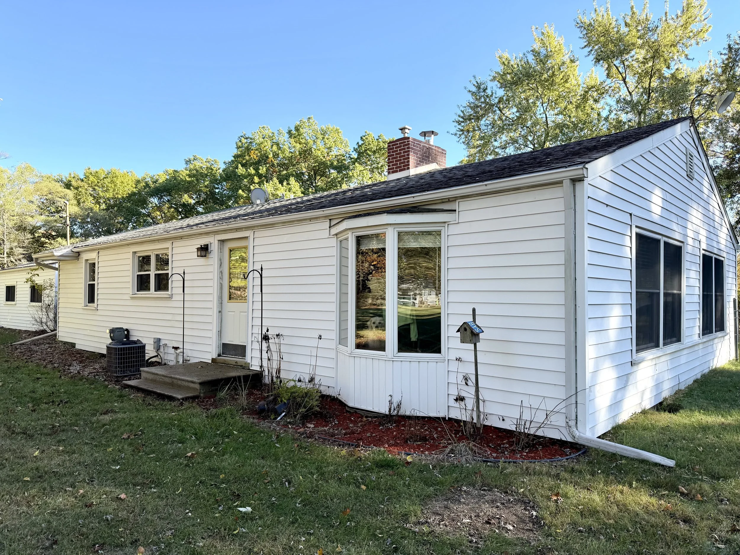 A white single-story house with siding, a brick chimney, and a sloped roof, surrounded by green trees and a grassy yard, with a small concrete porch and a black air conditioning unit outside.