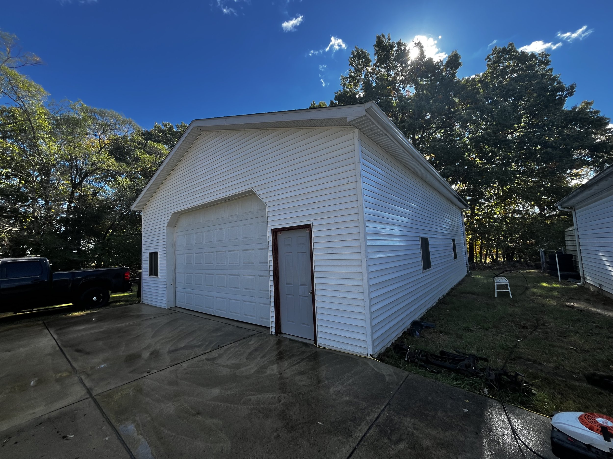 A white garage with a side door and small windows, set next to a house, with a wet concrete driveway, surrounded by green trees and a blue sky with some clouds.
