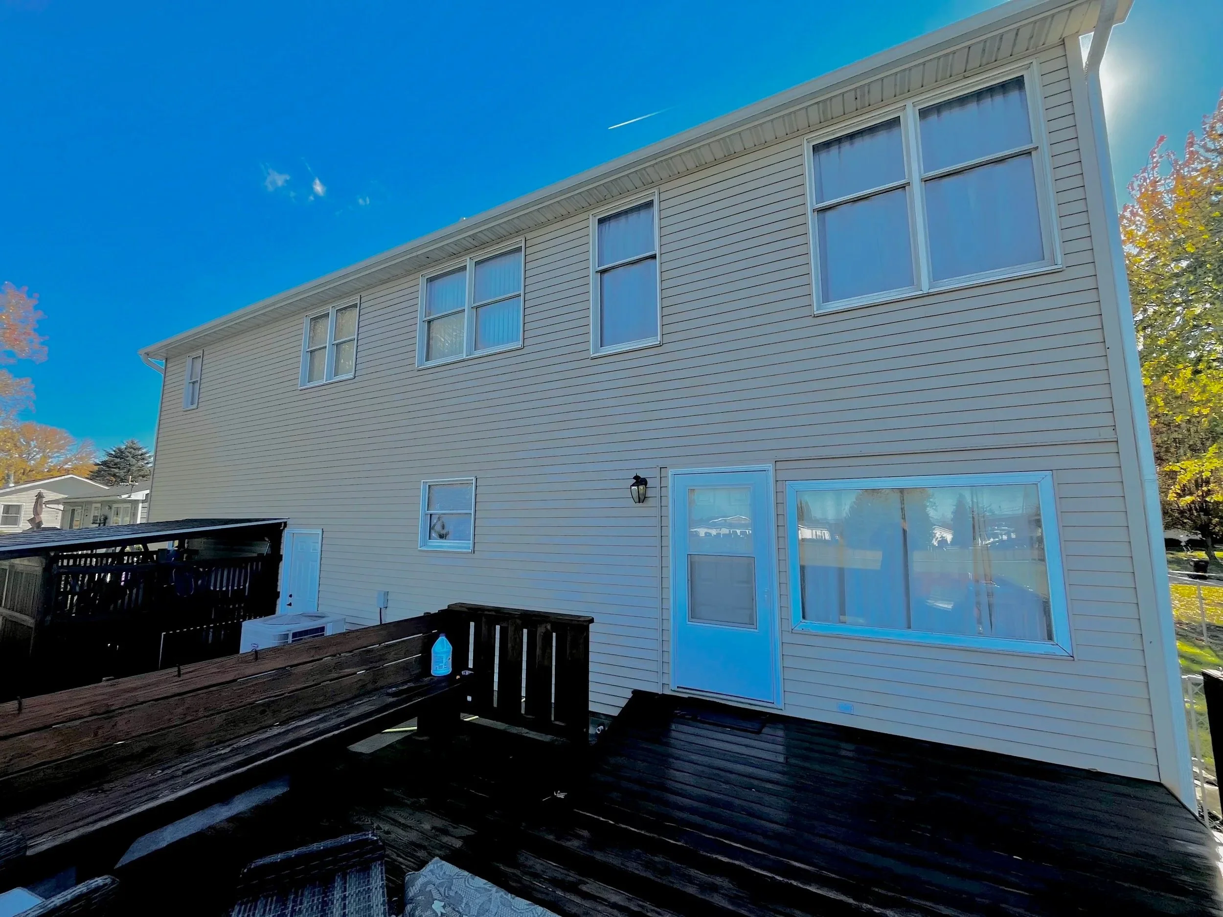 Back exterior of a two-story house with beige siding, multiple windows, a door with a large window, and a wooden deck with a cleaning spray bottle on the railing. The sky is clear with a jet trail visible.