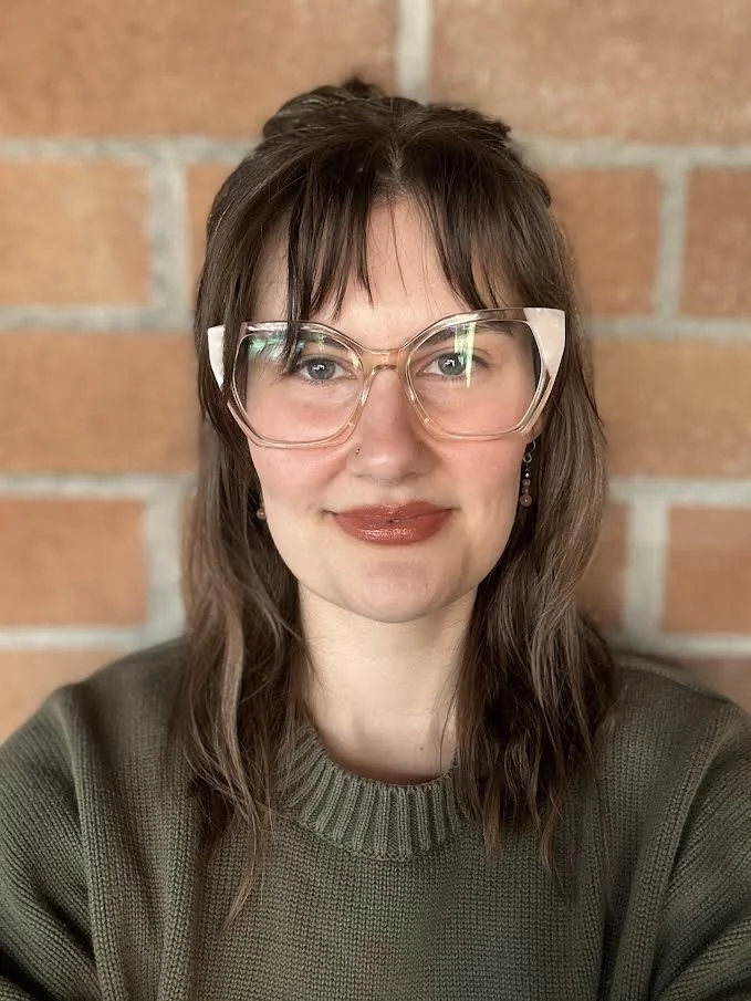 A woman with shoulder-length brown hair and glasses, smiling, standing in front of a brick wall.
