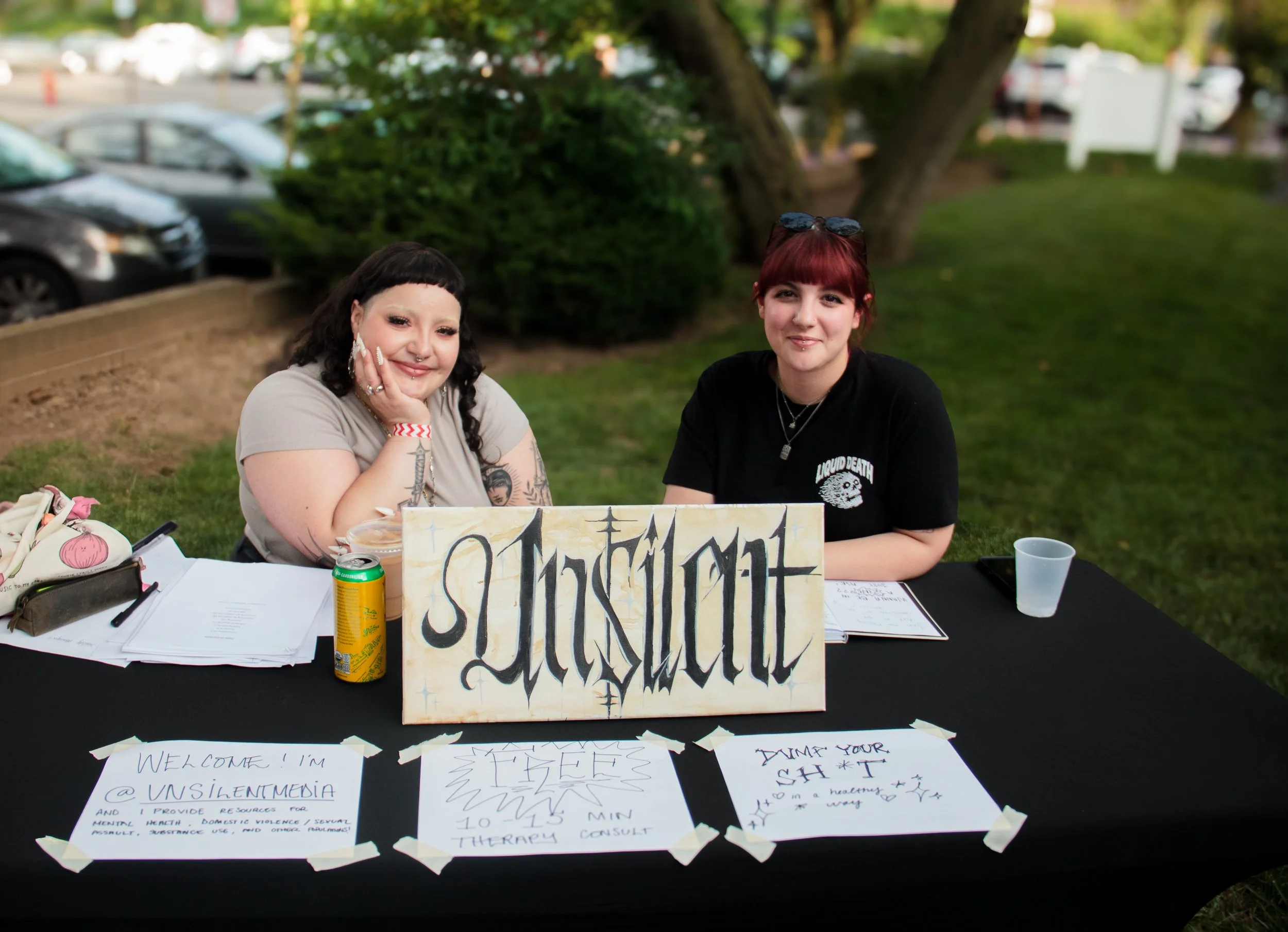 Two women sitting at a table outdoors with a sign that says "Insult" in stylized gothic font. The table has papers, a can of soda, and a cup. Behind them is green grass, trees, and parked cars.