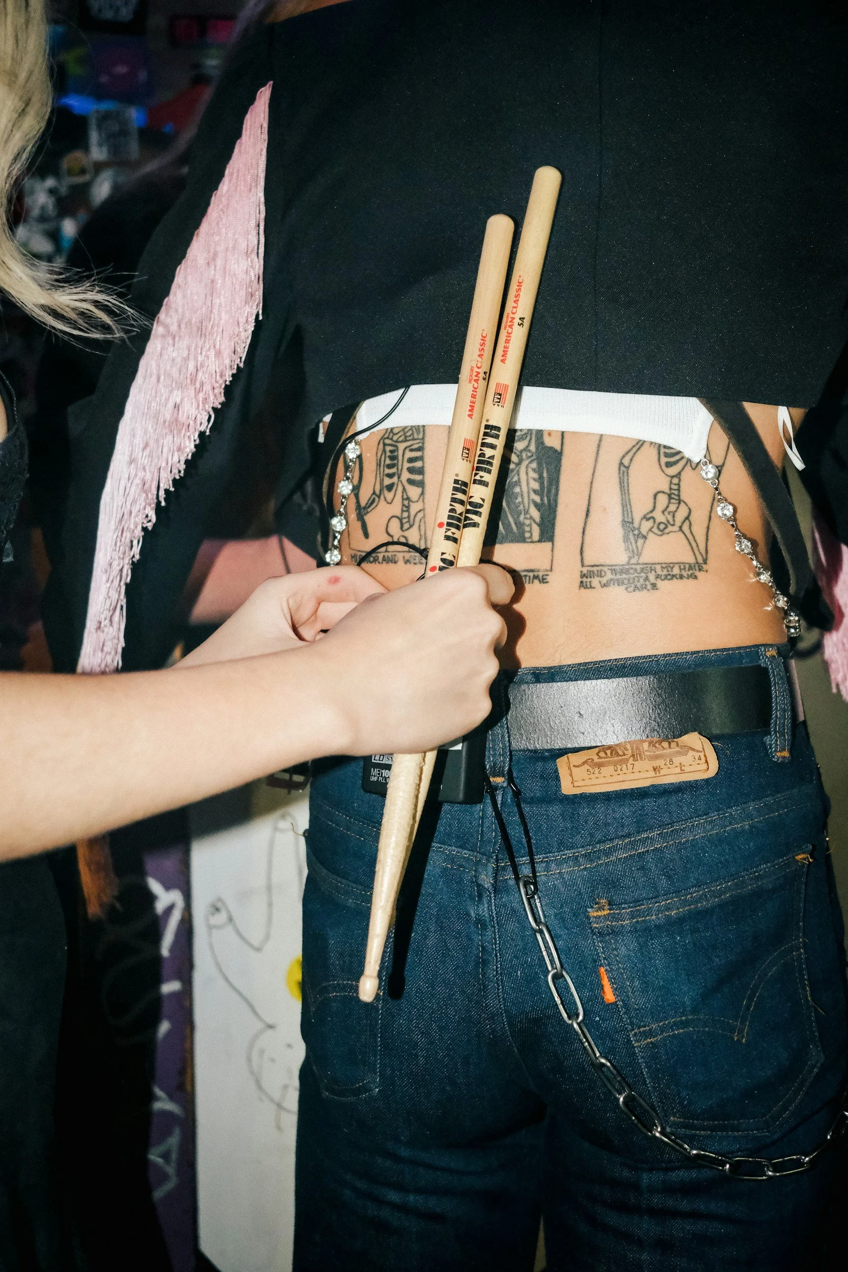 Person holding two wooden drumsticks behind their back, with tattoos on their back visible, wearing blue jeans and a black shirt with pink detail, in an indoor setting.