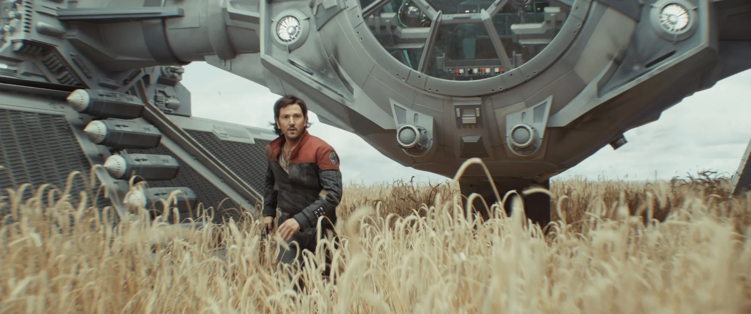 A man with dark hair and a beard wearing a black and brown jacket standing in a wheat field with a large spaceship behind him.