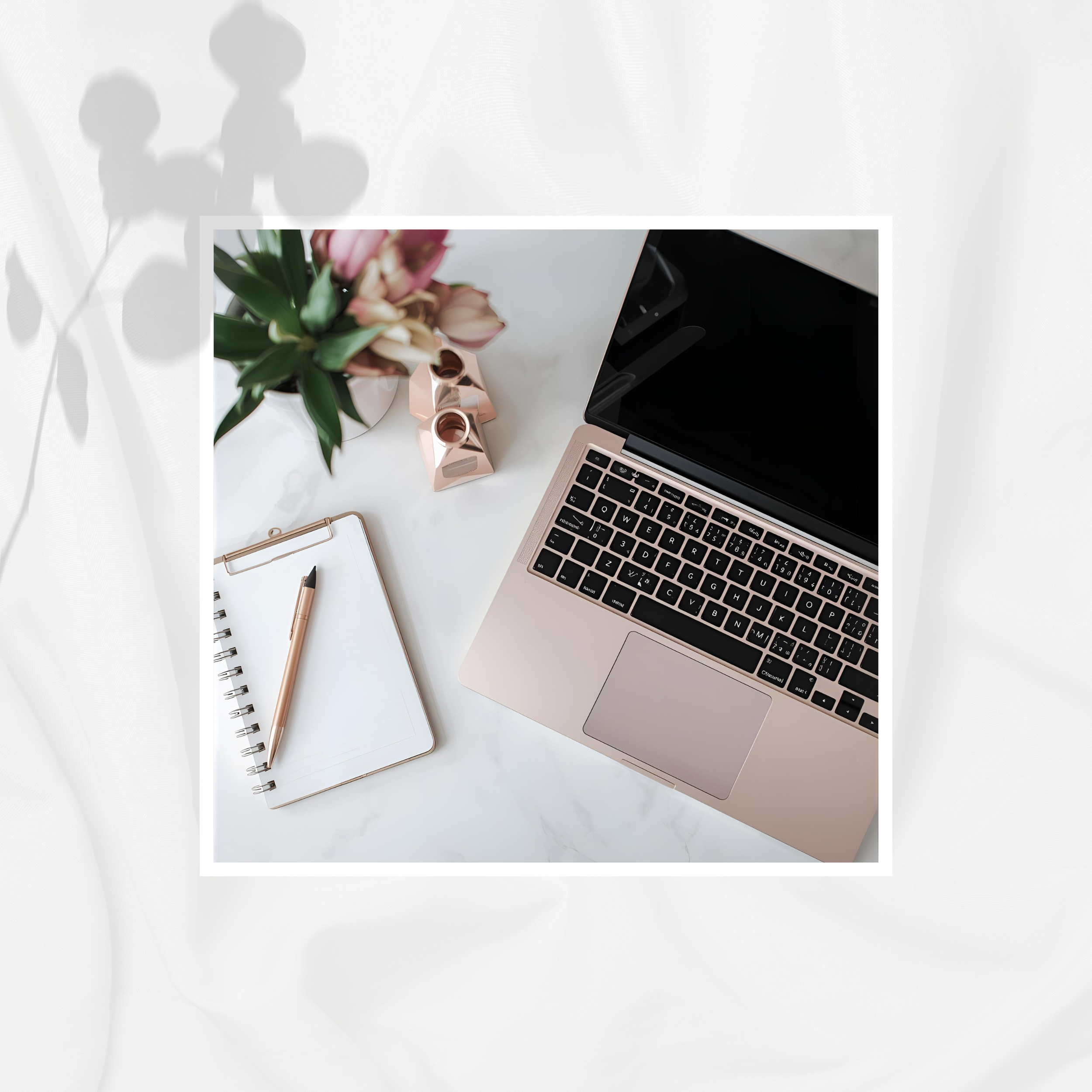 A laptop, notepad with pen, pink flowers in a pot, and two small pink decorative objects on a white marble desk.