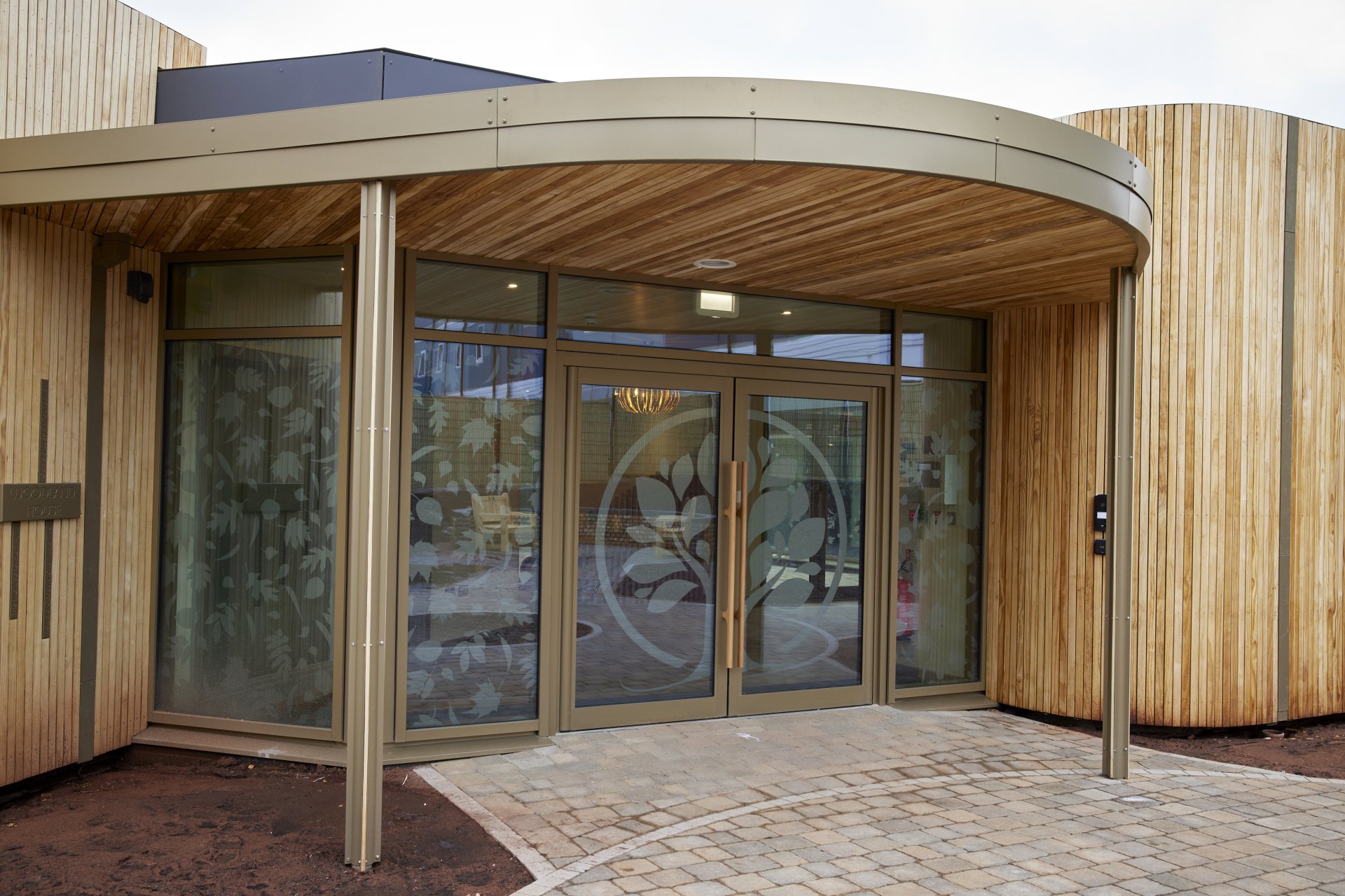 Modern building entrance with glass doors and wooden exterior, featuring a decorative tree design on the glass.