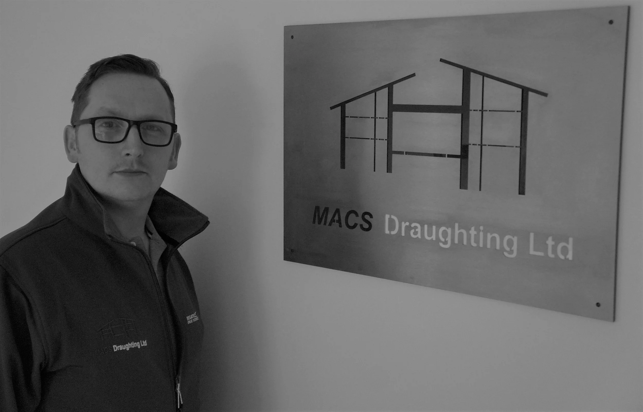 A man wearing glasses and a vest standing next to a sign that reads 'MACS Draughting Ltd' with a diagram of a building structure.